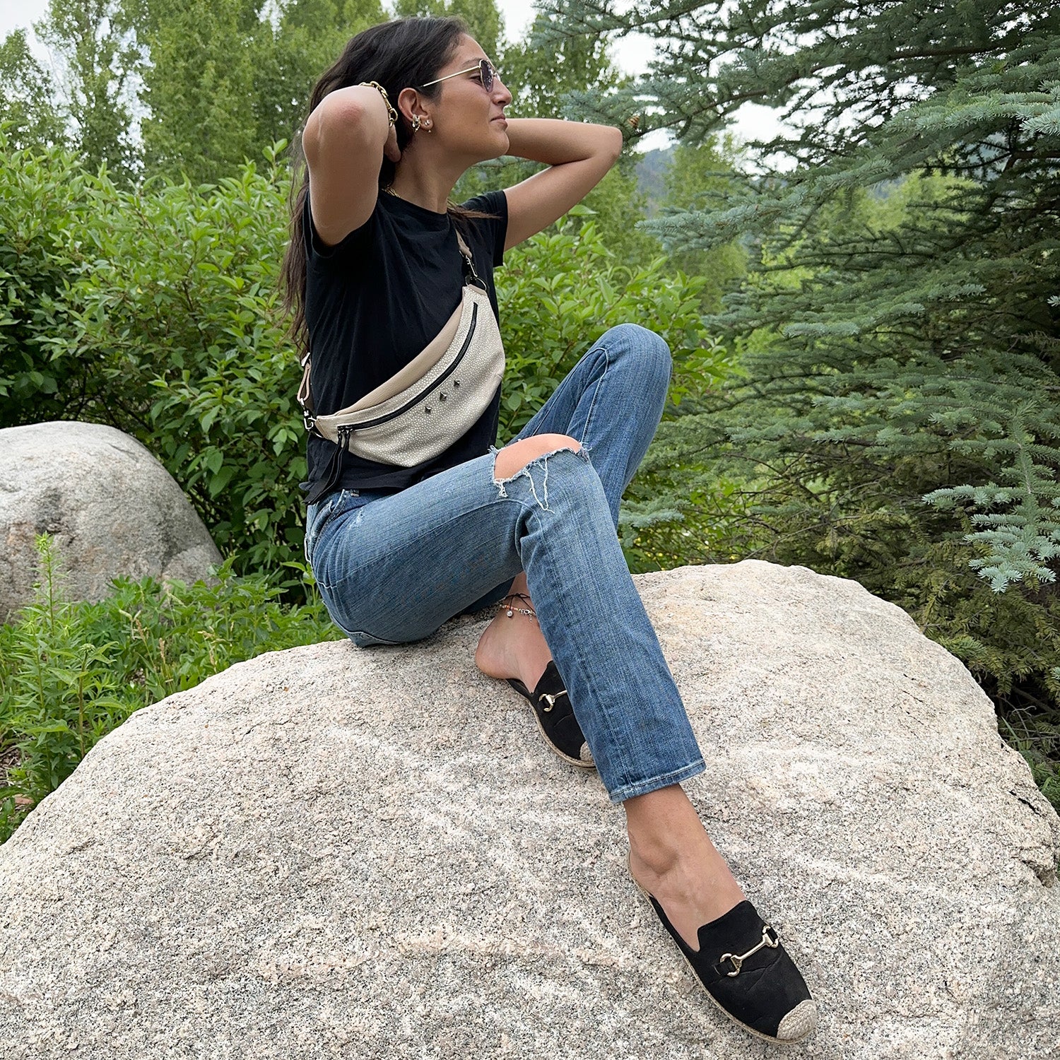 A person wearing sunglasses, a black shirt, jeans, and slip-on shoes sits on a rock amidst lush greenery, accompanied by the sleek STELLA - SAND STINGRAY crossbody bag by evokewomen.