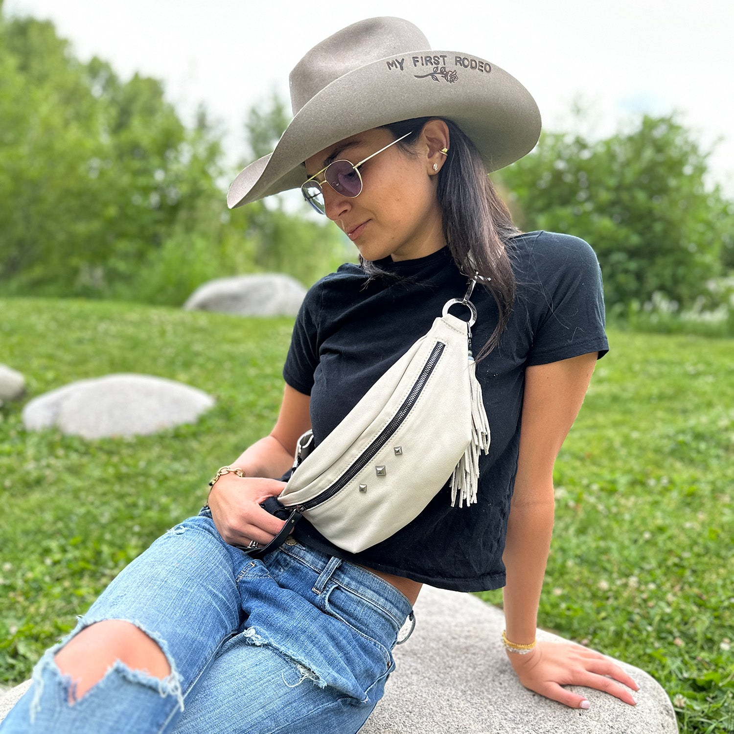 A woman in a cowboy hat and sunglasses poses outdoors wearing a black t-shirt, ripped jeans, and the STELLA - PUTTY beige fringe crossbody bag by evokewomen.