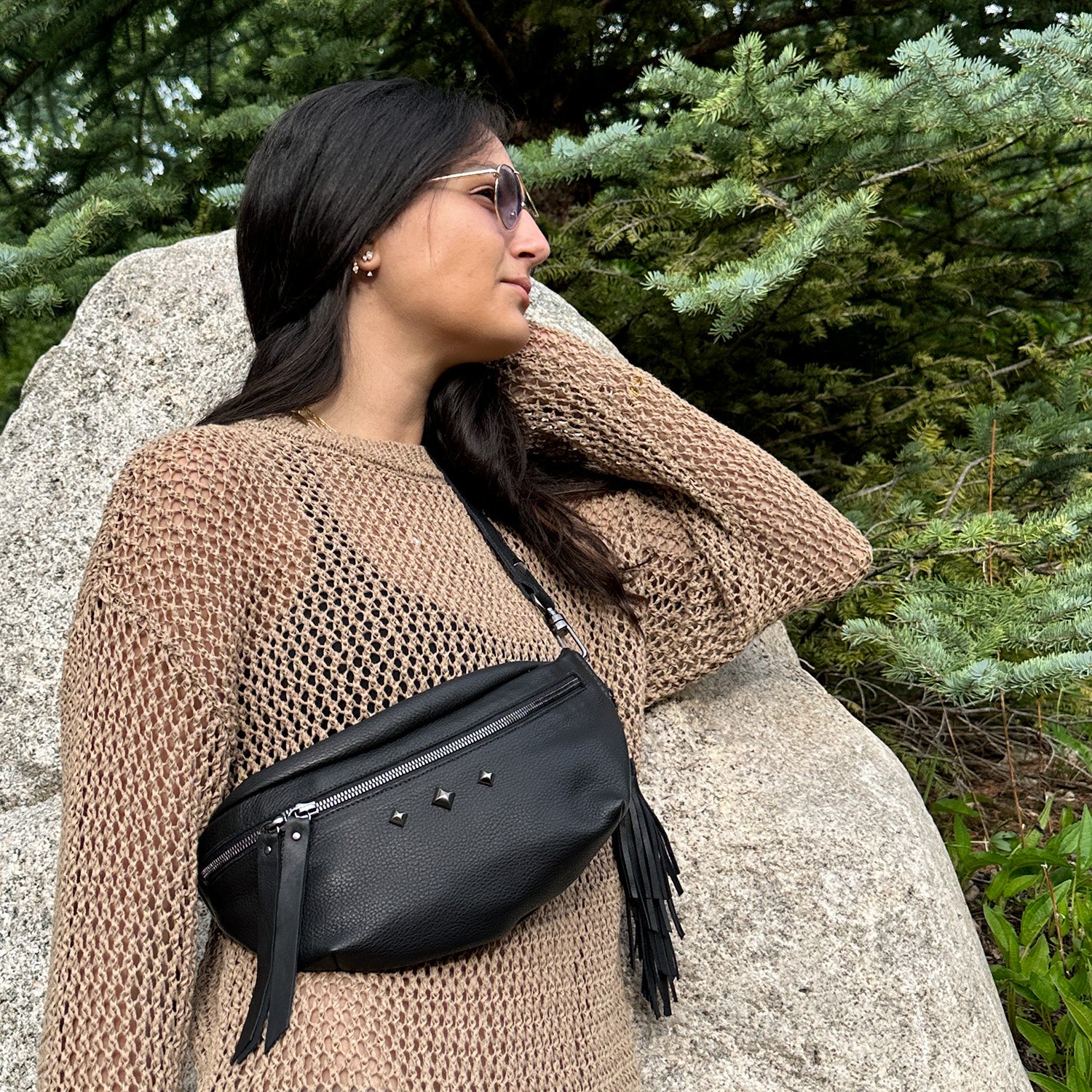 A woman in sunglasses and a brown sweater stands by a large rock, with the STELLA - BLACK crossbody bag from evokewomen slung across her shoulder. She is outdoors, surrounded by green trees.
