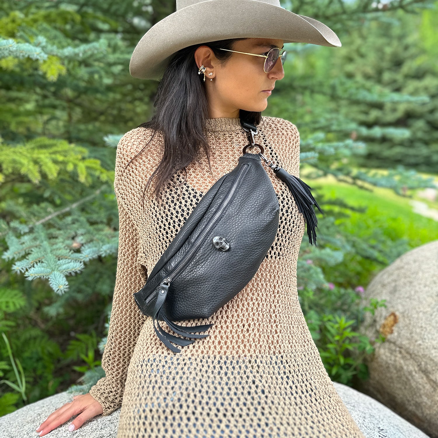 A woman in a wide-brimmed hat and sunglasses stands outdoors wearing a beige mesh dress and the STELLA - BLACK stylish leather sling bag by evokewomen.