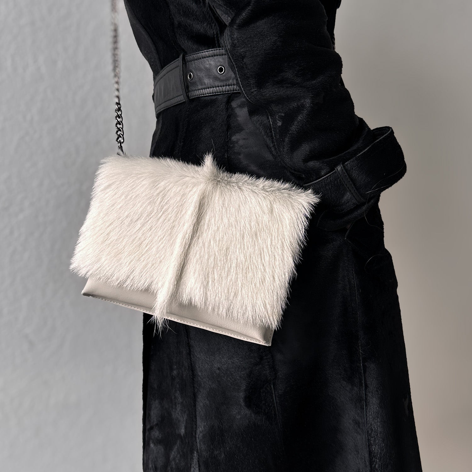 A person wearing a black coat holds the evokewomen BAHAMA FUR - WINTER WHITE bag, a luxurious and versatile accessory that effortlessly elevates any outfit.