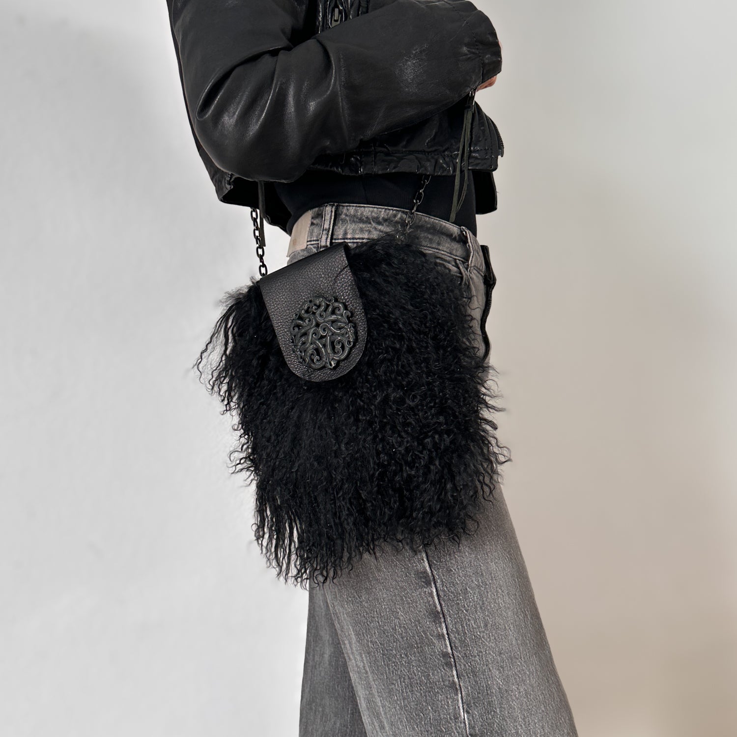 A person dressed in a black jacket and gray jeans, accessorized with the stylish evokewomen MIA - BLACK crossbody bag made from Mongolian lamb fur.
