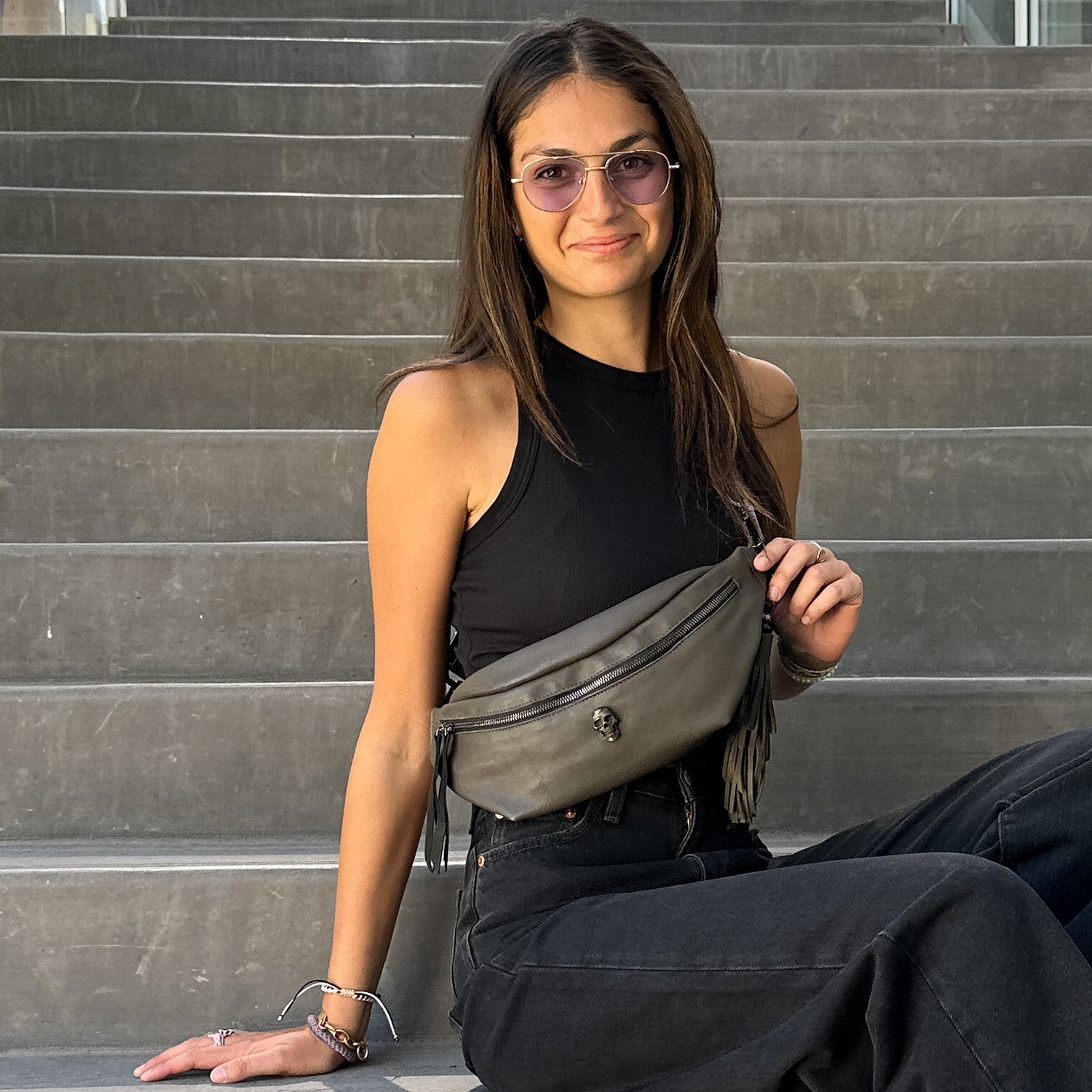 A woman wearing sunglasses, a black sleeveless top, and dark pants sits on concrete steps with the STELLA - OLIVE-GREY sling bag by evokewomen gracefully draped across her.