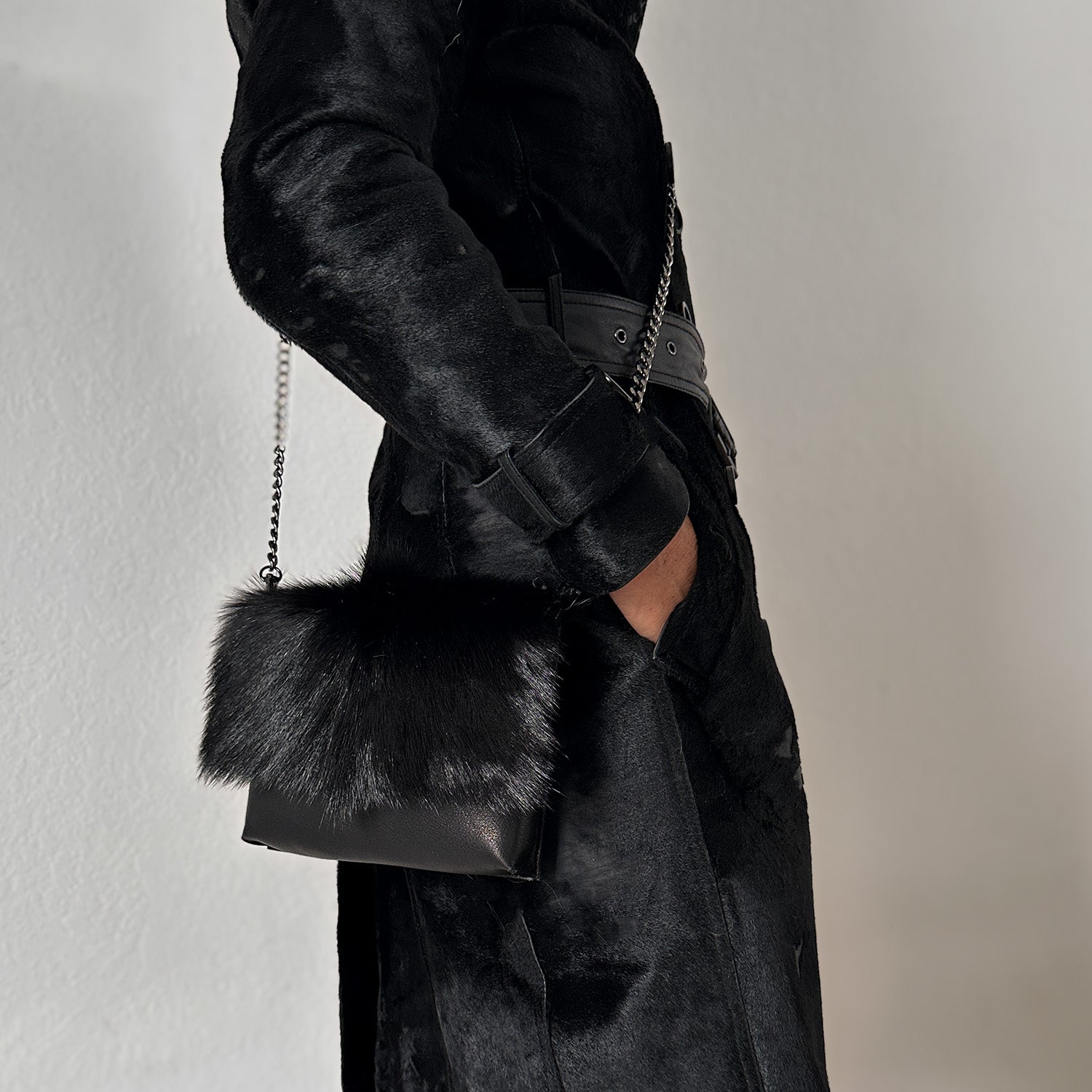 A person in an opulent black fur coat holds the luxurious BAHAMA FUR - BLACK bag from evokewomen, featuring a sleek chain strap.