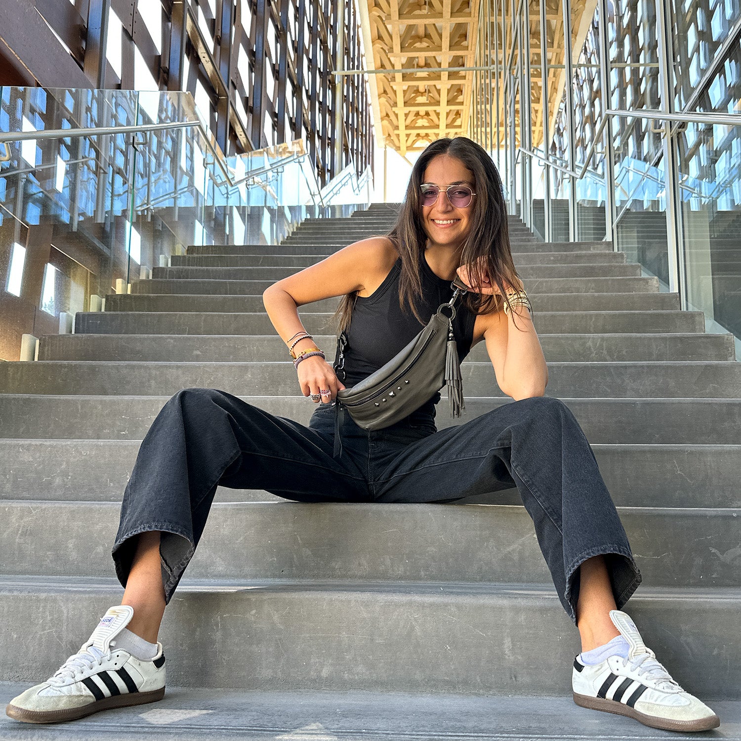 A person is seated on broad concrete steps beneath a modern architectural canopy, sporting sunglasses, a black top, dark jeans, and white sneakers. They have the sleek STELLA - OLIVE-GREY sling bag from evokewomen draped across their shoulder with its adjustable strap.