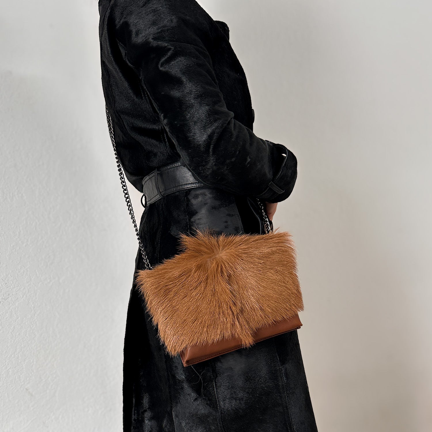 A person in a black coat carries the chic BAHAMA FUR - COGNAC handbag by evokewomen, featuring a chain strap and styled as a crossbody bag.