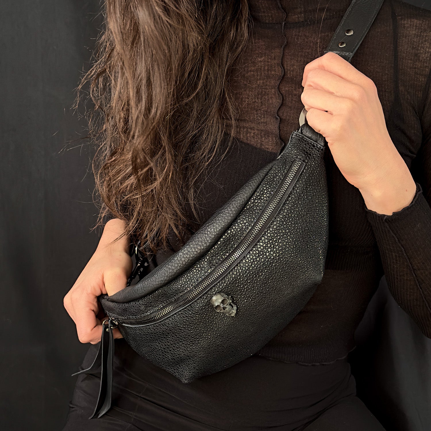 A person in a black top holds the sleek STELLA - BLACK STINGRAY crossbody bag from evokewomen, featuring a zipper pocket and small skull detail.