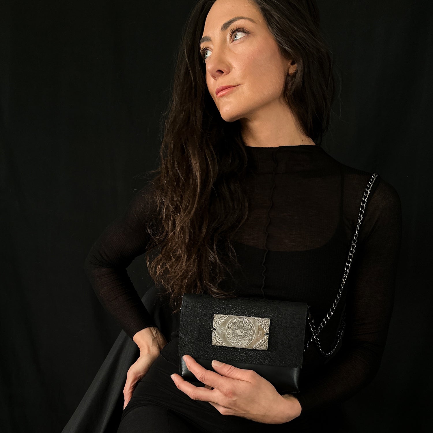 A person dressed in black is holding the BAHAMA - BLACK STINGRAY bag by evokewomen, featuring a silver emblem and a crossbody chain, set against a dark background.