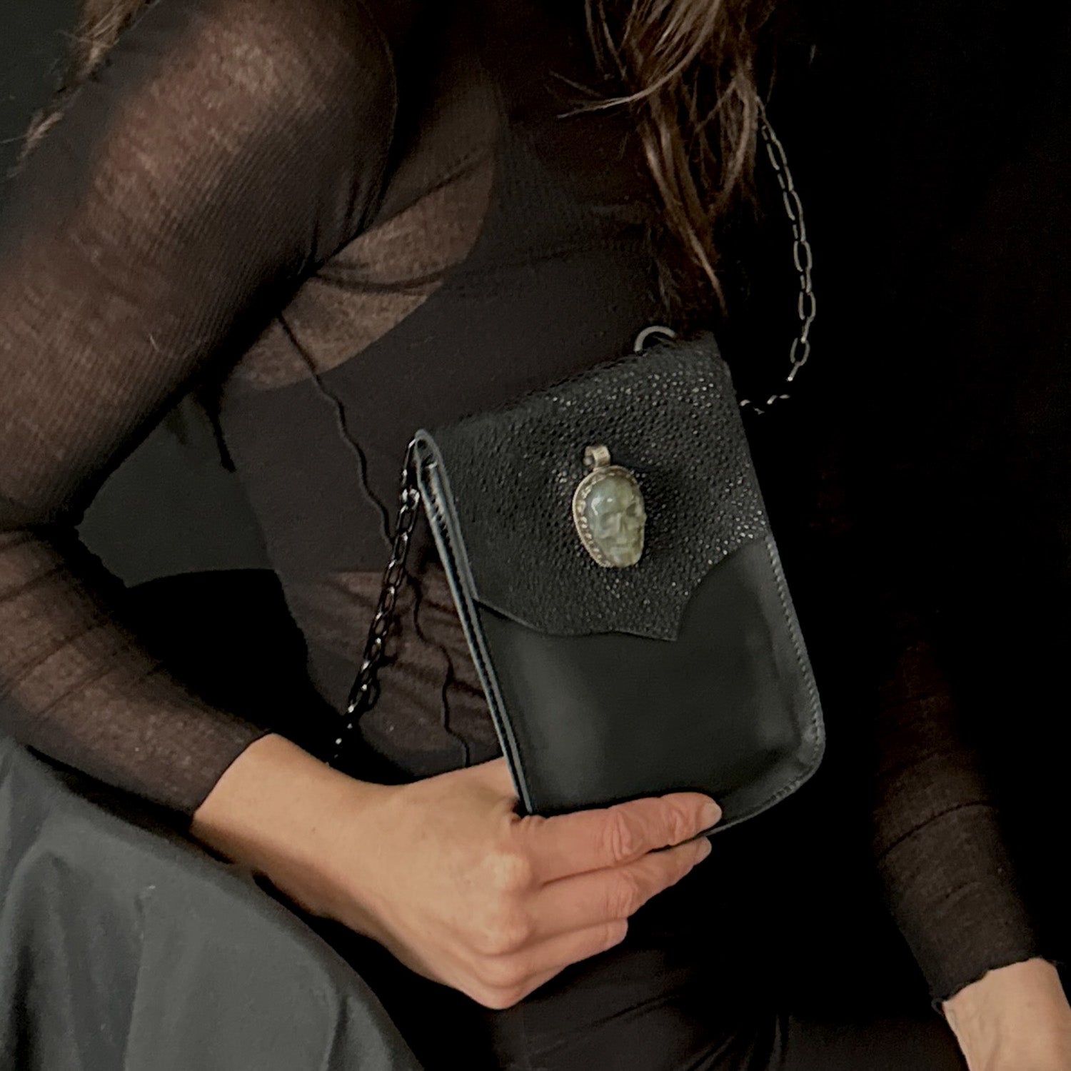 A person carries the PAULINA -BLACK STINGRAY crossbody bag by evokewomen, crafted from exquisite Italian leather and adorned with a unique labradorite skull amulet on the front.