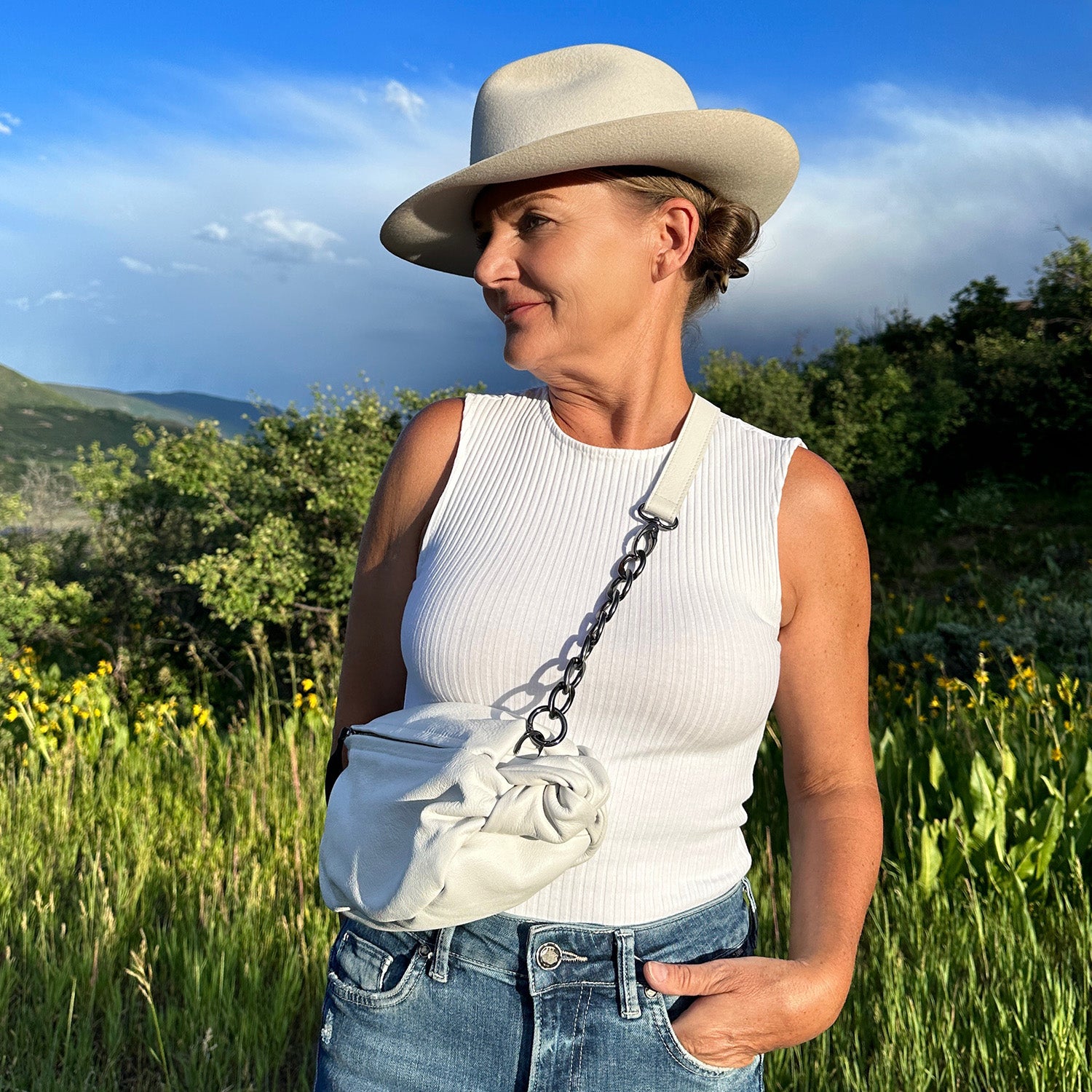 A woman wearing the LONDON - WHITE white hat and tank top from evokewomen stands outdoors, showcasing a stylish crossbody bag against a backdrop of lush greenery and a clear sky.