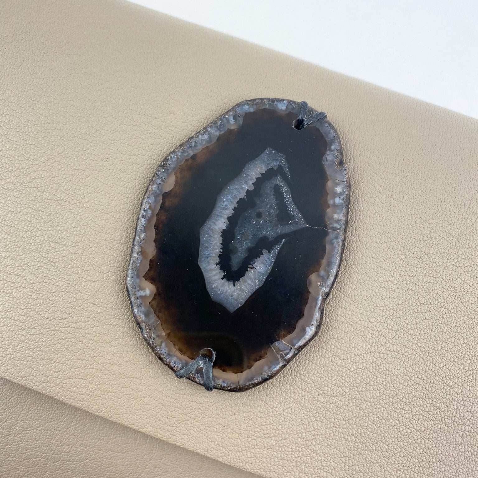 An oval-shaped geode slice with a dark center and lighter edges is carefully placed on the beige leather of an evokewomen BAHAMA - SAND bag.