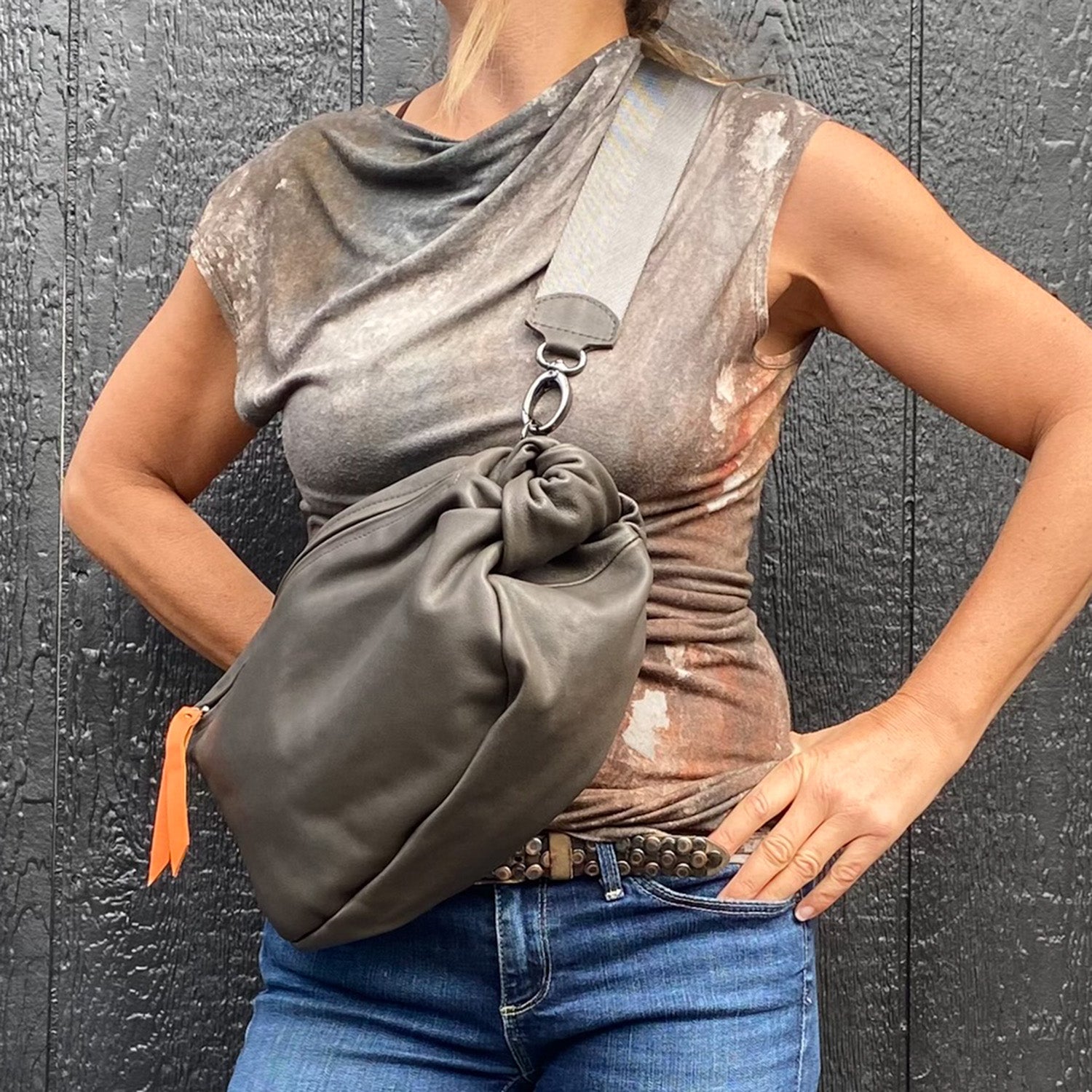 A person wearing a tie-dye top and jeans holds the LULA - OLIVE-GREY sling bag with an orange leather puller from evokewomen, standing in front of a black textured wall.