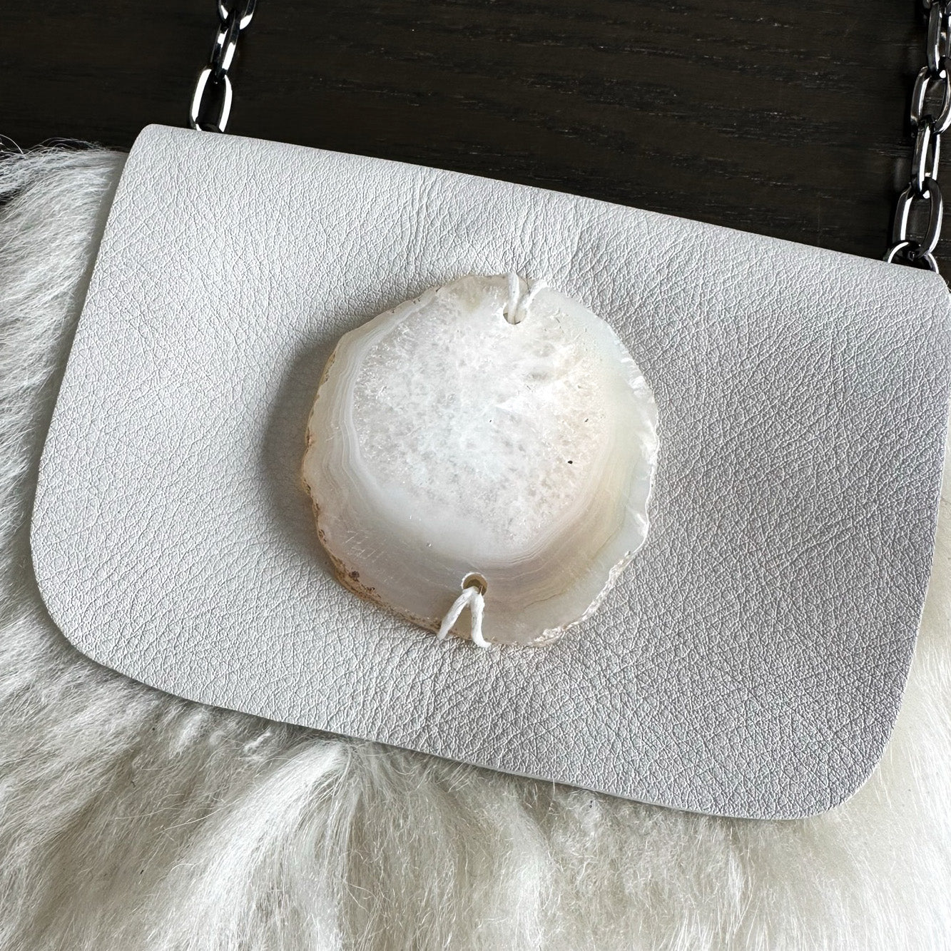 The MIA - OFF WHITE by evokewomen is a white leather crossbody bag featuring a large round agate stone centered on the flap, elegantly displayed on a furry white surface.