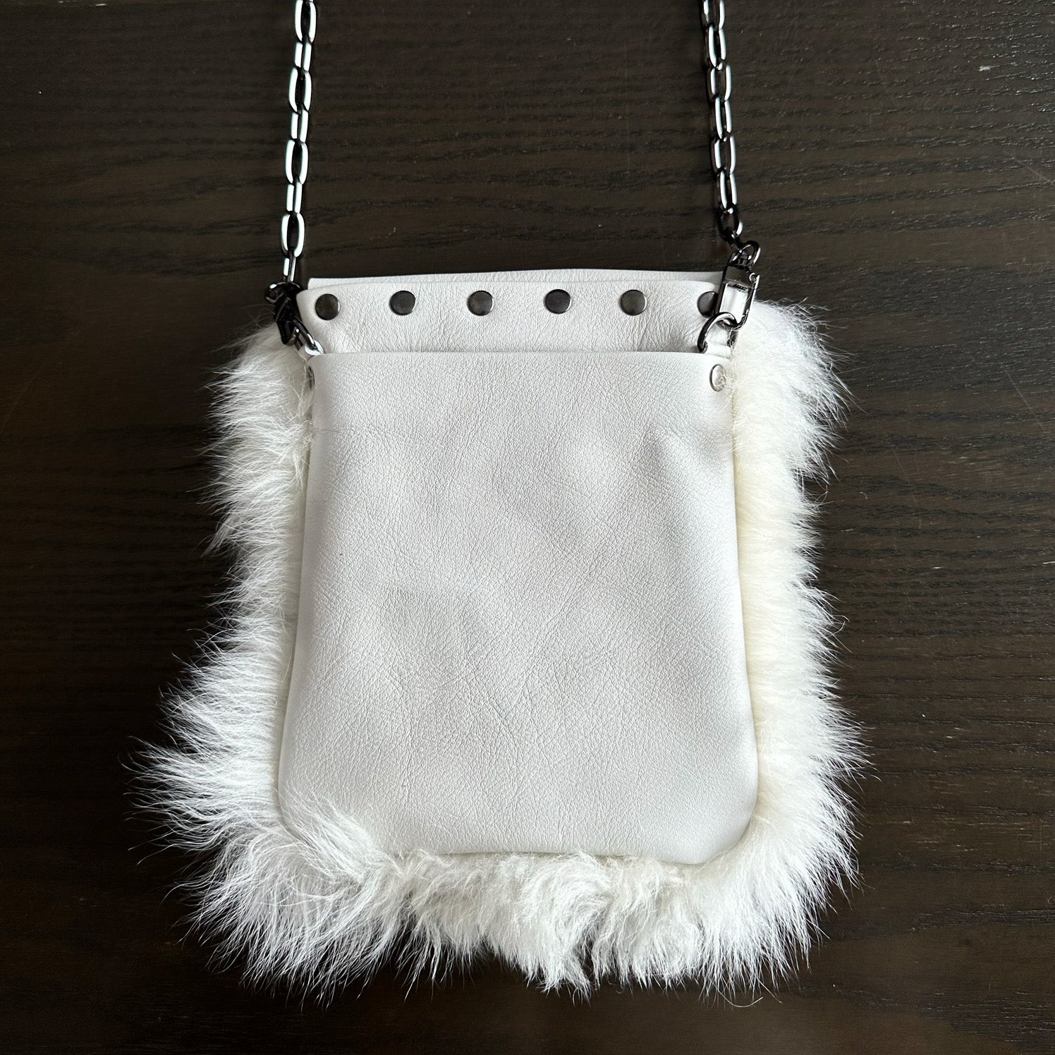 The MIA - OFF WHITE bag by evokewomen, featuring a chic white leather design with faux fur trim and a chain strap, rests elegantly on a dark wood surface.