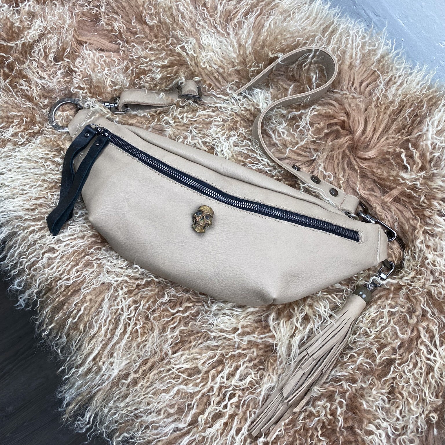 The STELLA - SAND by evokewomen, a beige leather fanny pack featuring a skull emblem, adjustable strap, black zipper, and tassel, is displayed on a fluffy surface.