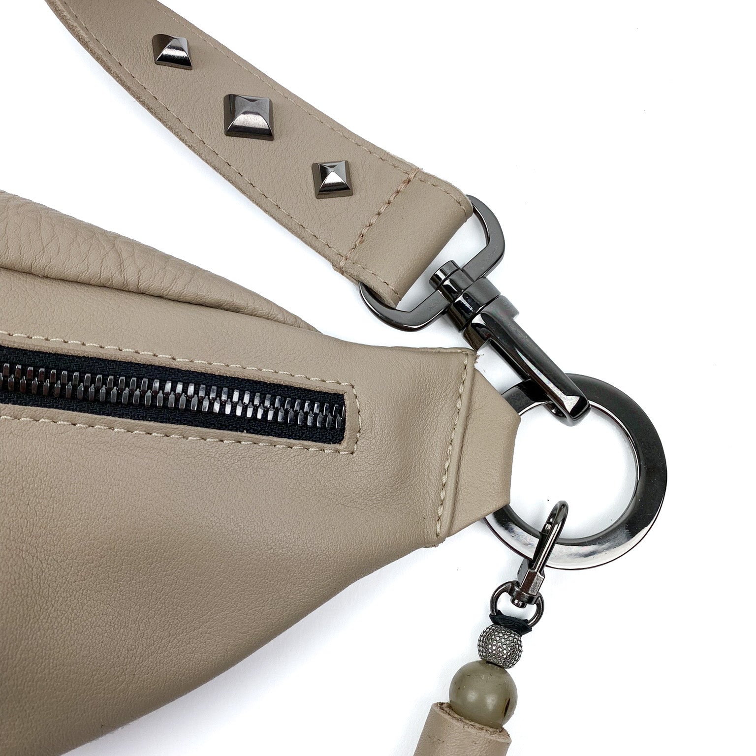 Close-up of the STELLA - SAND sling bag by evokewomen, featuring beige leather with a metal zipper and a strap adorned with silver studs. The crossbody strap comes with a silver clasp and includes a decorative keychain with beads, adding an elegant touch.