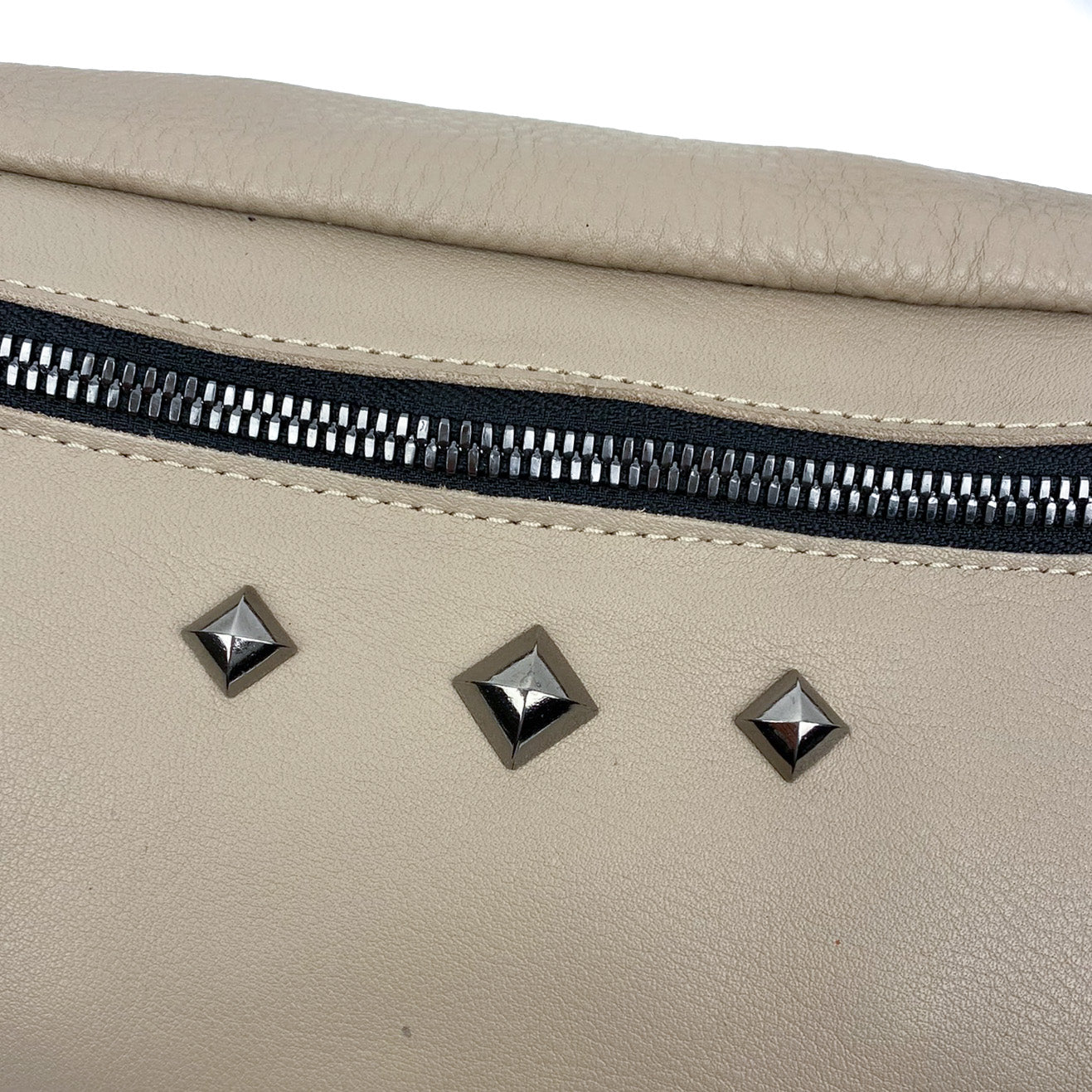 Close-up of the STELLA - SAND cross body bag by evokewomen, featuring beige leather, a black zipper, and three metallic diamond-shaped studs.