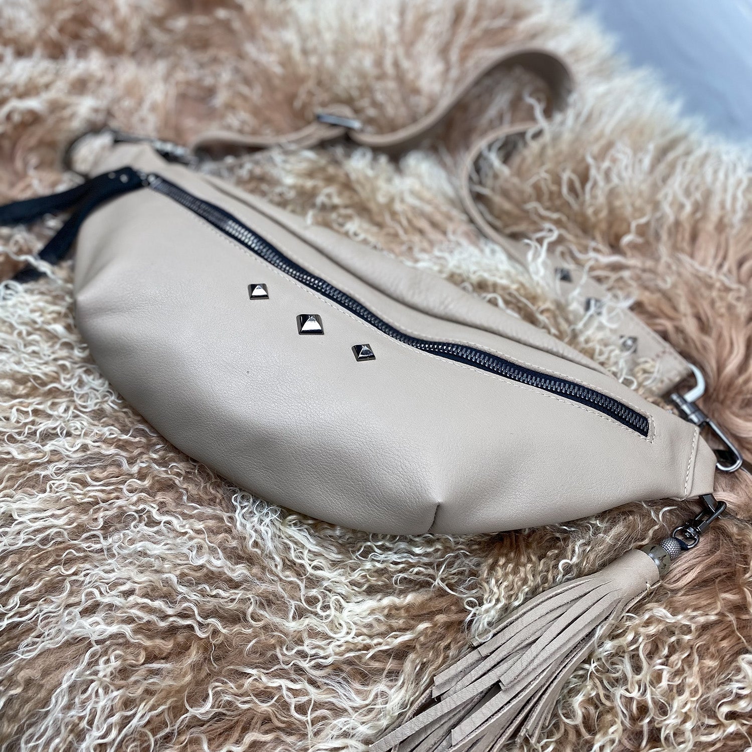The STELLA - SAND waist bag by evokewomen, crafted from beige leather and adorned with a zipper and decorative studs, rests on a fluffy, textured surface. A tassel is elegantly attached to this sleek accessory.