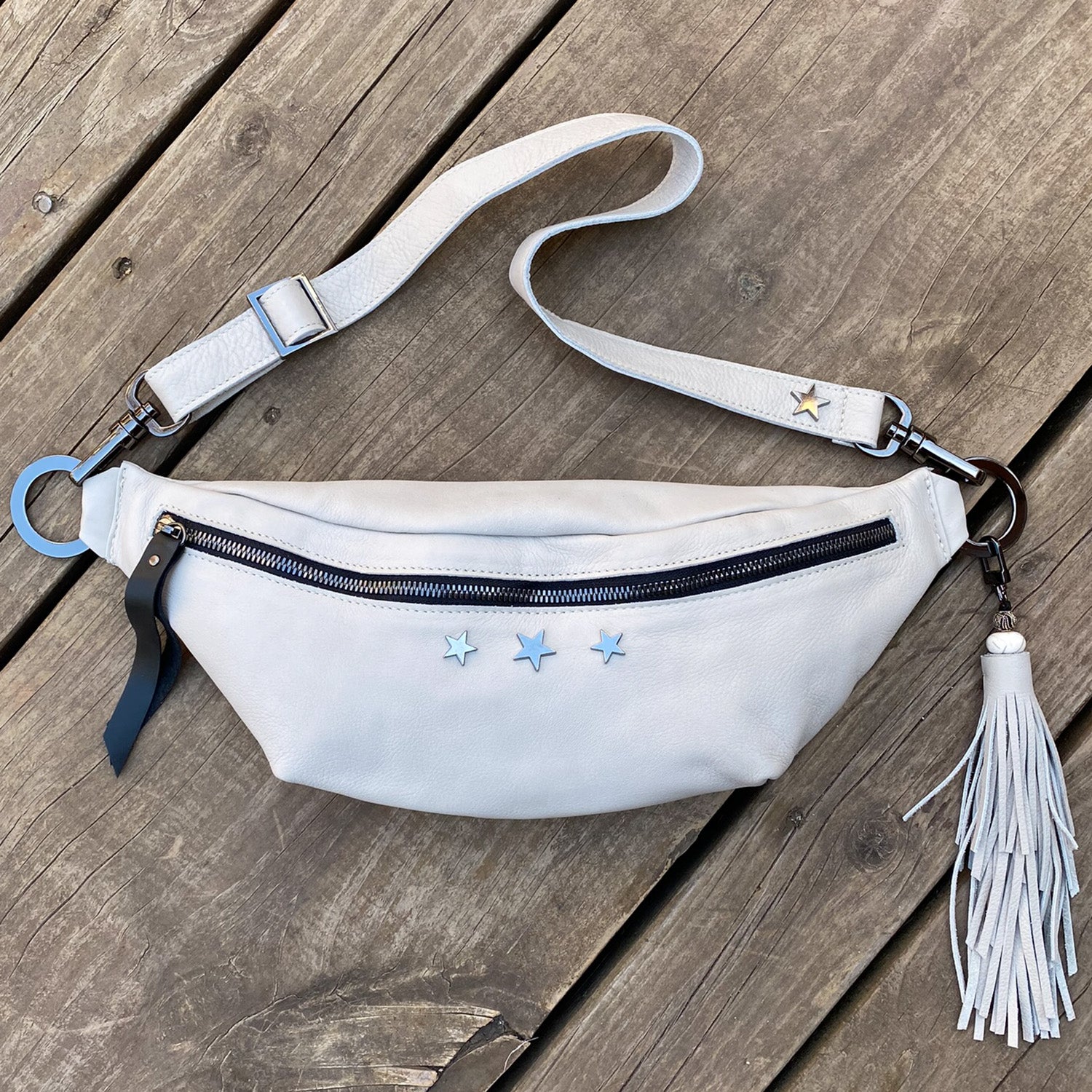 Displayed on a wooden surface, the STELLA - PUTTY by evokewomen is a light gray leather crossbody bag adorned with a front zipper, star decorations, and a tassel, providing the stylish versatility of a sling bag.
