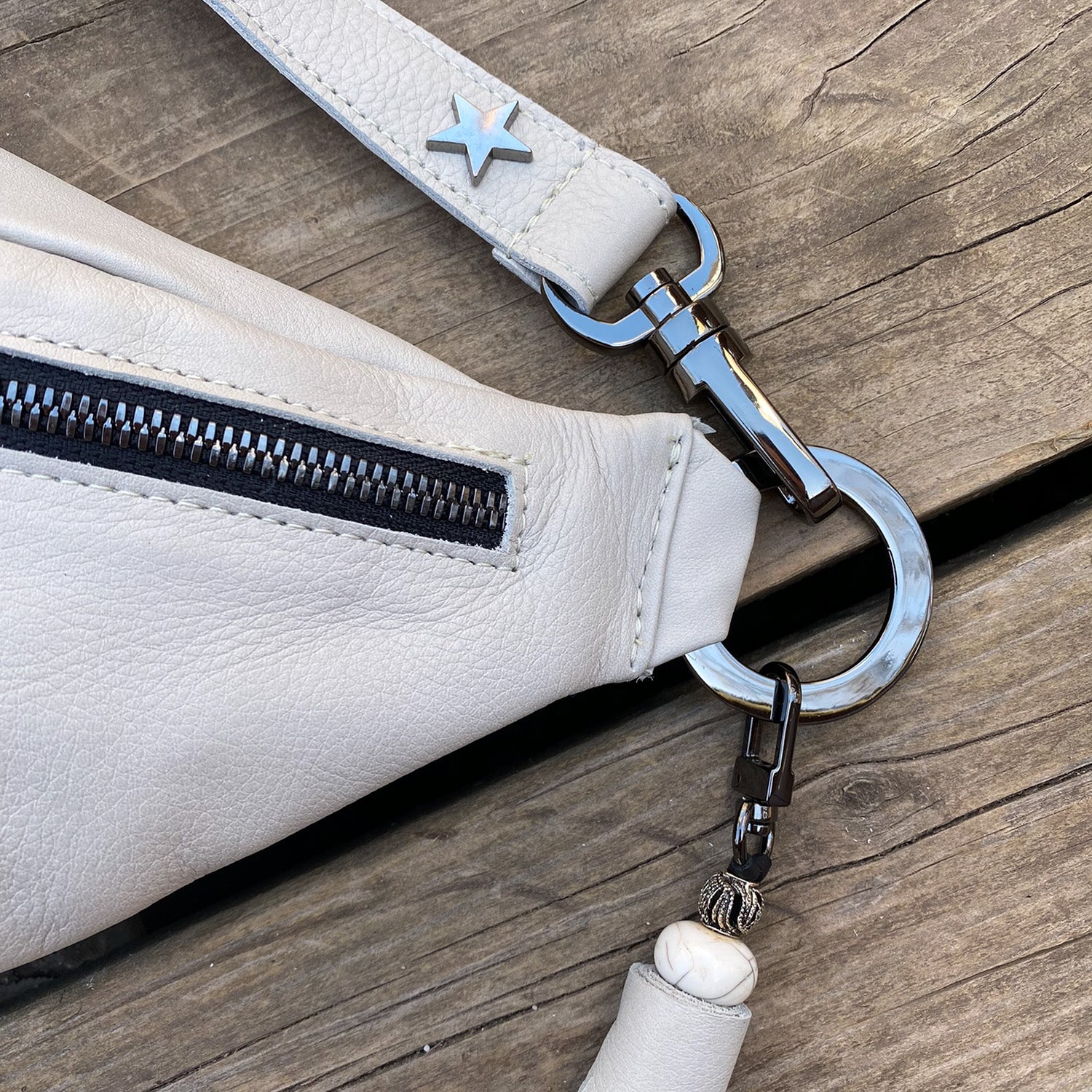 Close-up of the evokewomen STELLA - PUTTY, a light gray leather crossbody bag featuring a black zipper, metal buckle, and star decoration on the strap, placed on a wooden surface.