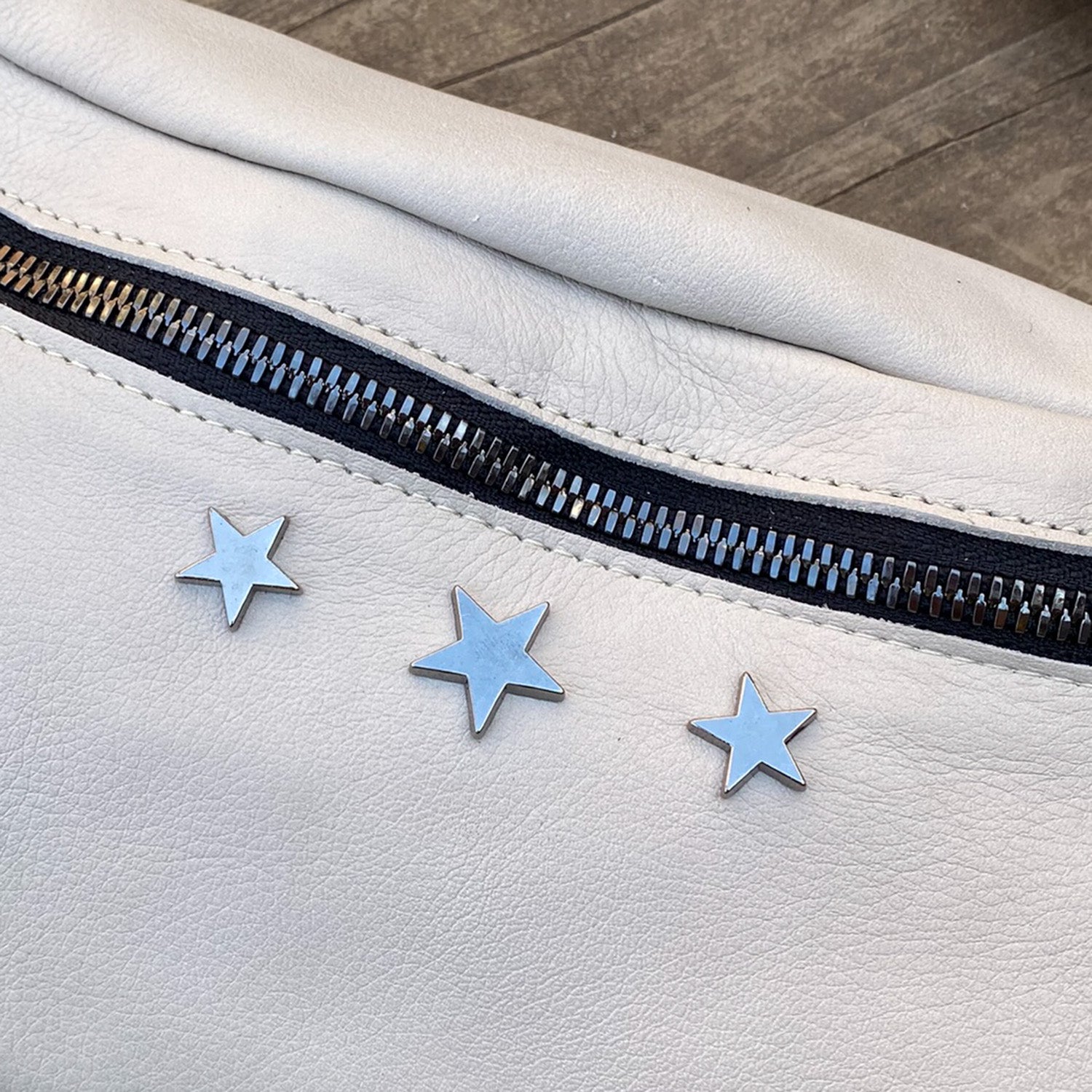 The STELLA - PUTTY by evokewomen is a white leather sling bag featuring a black zipper and three metallic star-shaped studs on the surface.
