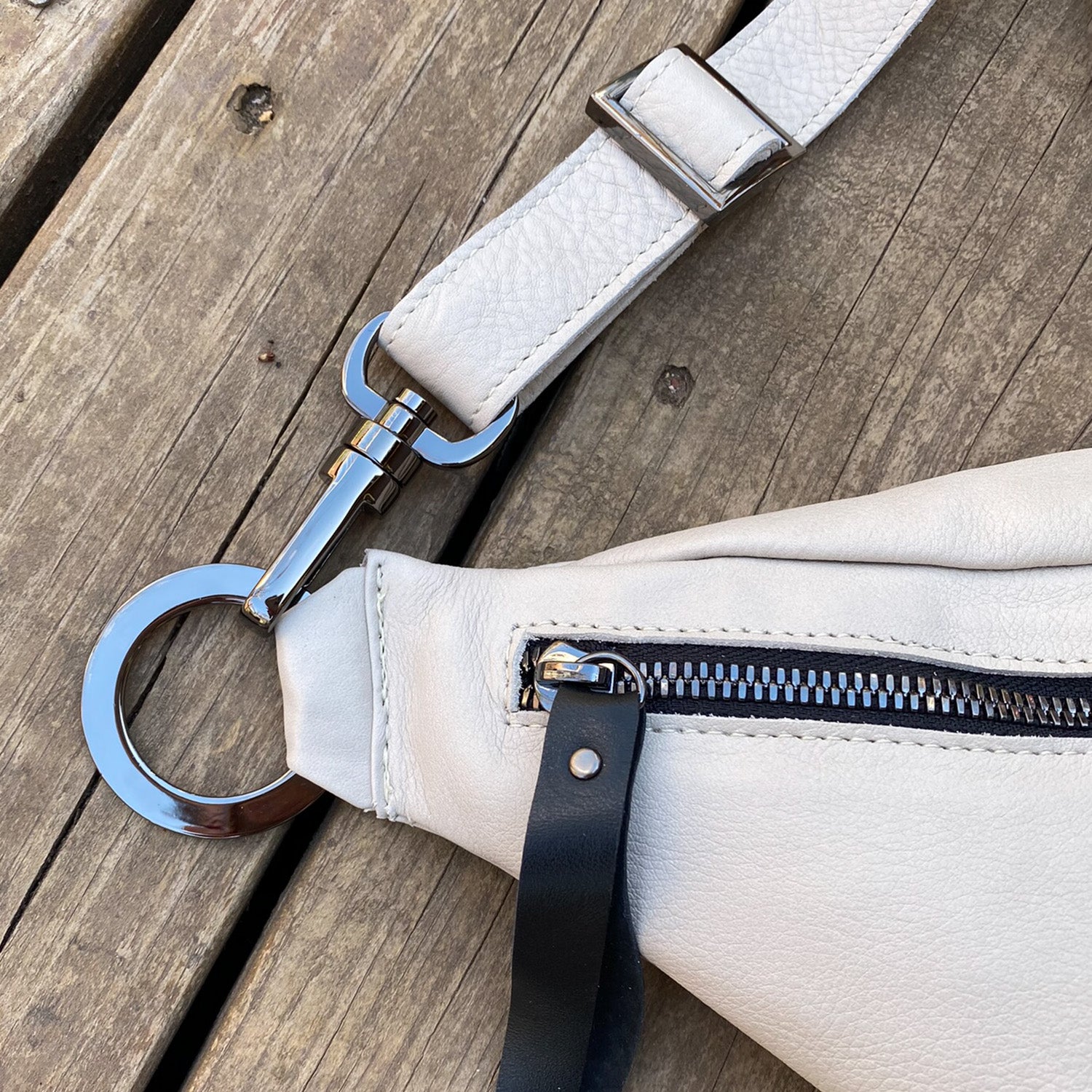 Close-up of the STELLA - PUTTY white leather cross body bag by evokewomen, featuring a zipper pocket and a metal clasp attachment, resting on a wooden surface.