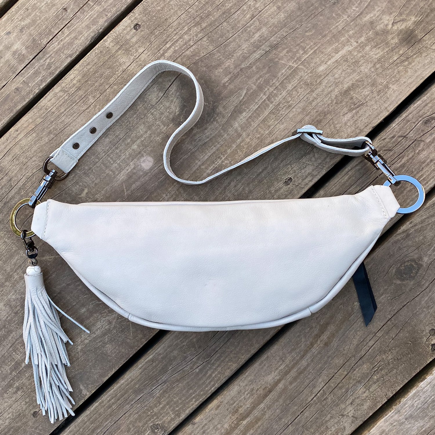 The STELLA - PUTTY by evokewomen, a white crescent-shaped cross body bag featuring a strap and fringe tassel, rests elegantly on a wooden surface.