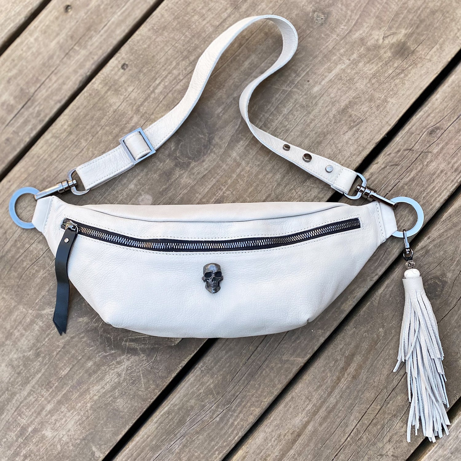 The STELLA - PUTTY by evokewomen is a white leather crossbody bag featuring a zipper, skull embellishment, and fringe tassel, beautifully displayed on a wooden surface.