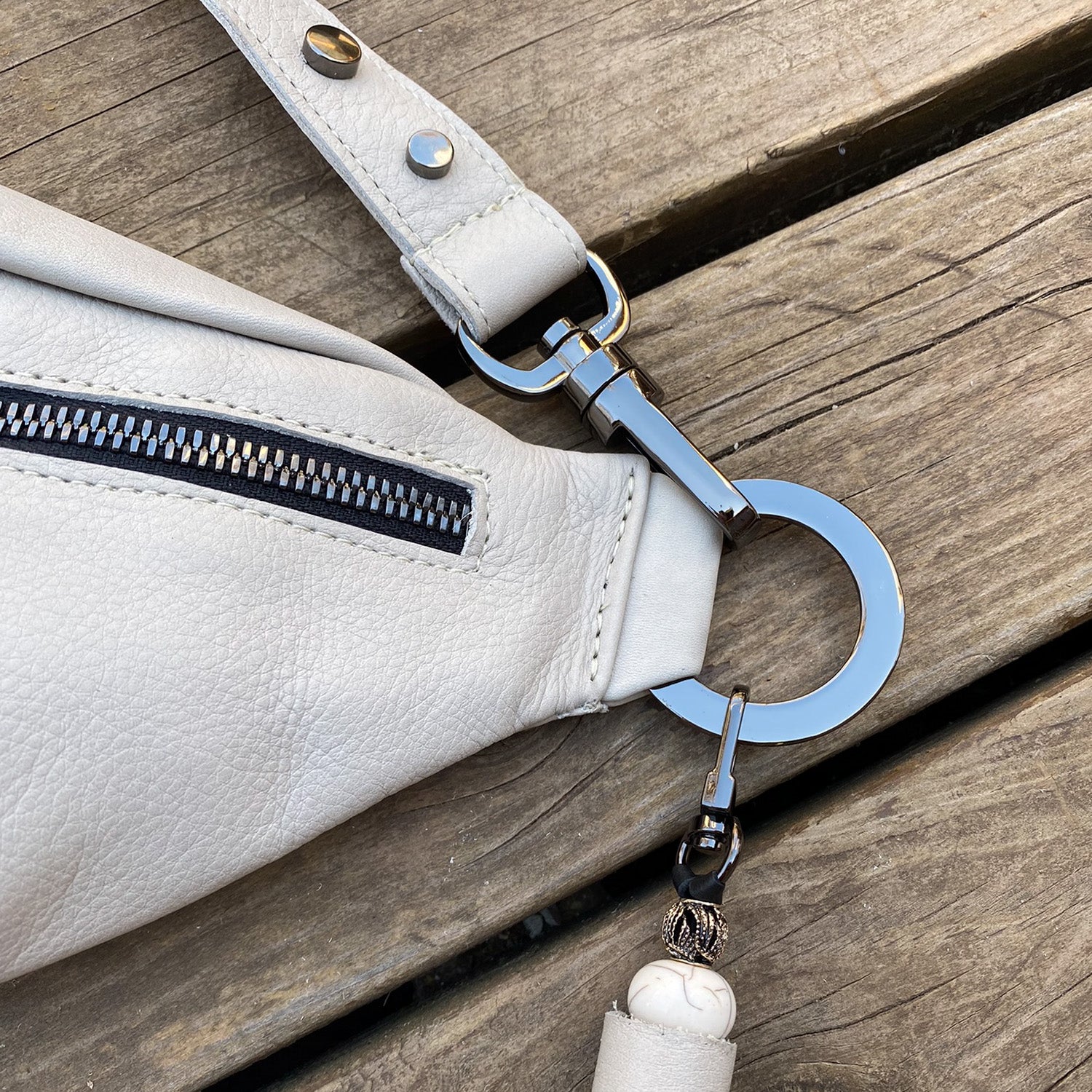 Close-up of the STELLA - PUTTY, a white leather cross body bag by evokewomen, featuring a metal zipper on a wooden surface. A metal ring and clasp with a decorative bead are attached to the bag's strap, adding an elegant touch to this versatile accessory.