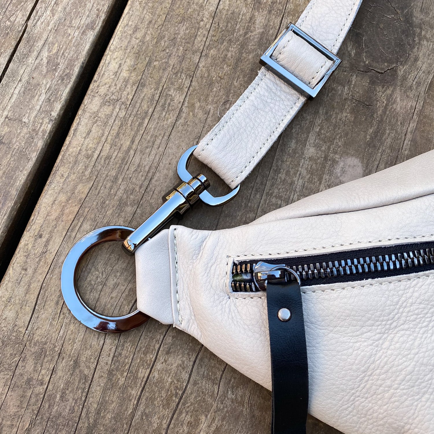 Close-up of the STELLA - PUTTY crossbody bag by evokewomen, showcasing its beige leather design with a metal ring, zipper, and adjustable shoulder strap, placed on a wooden surface.