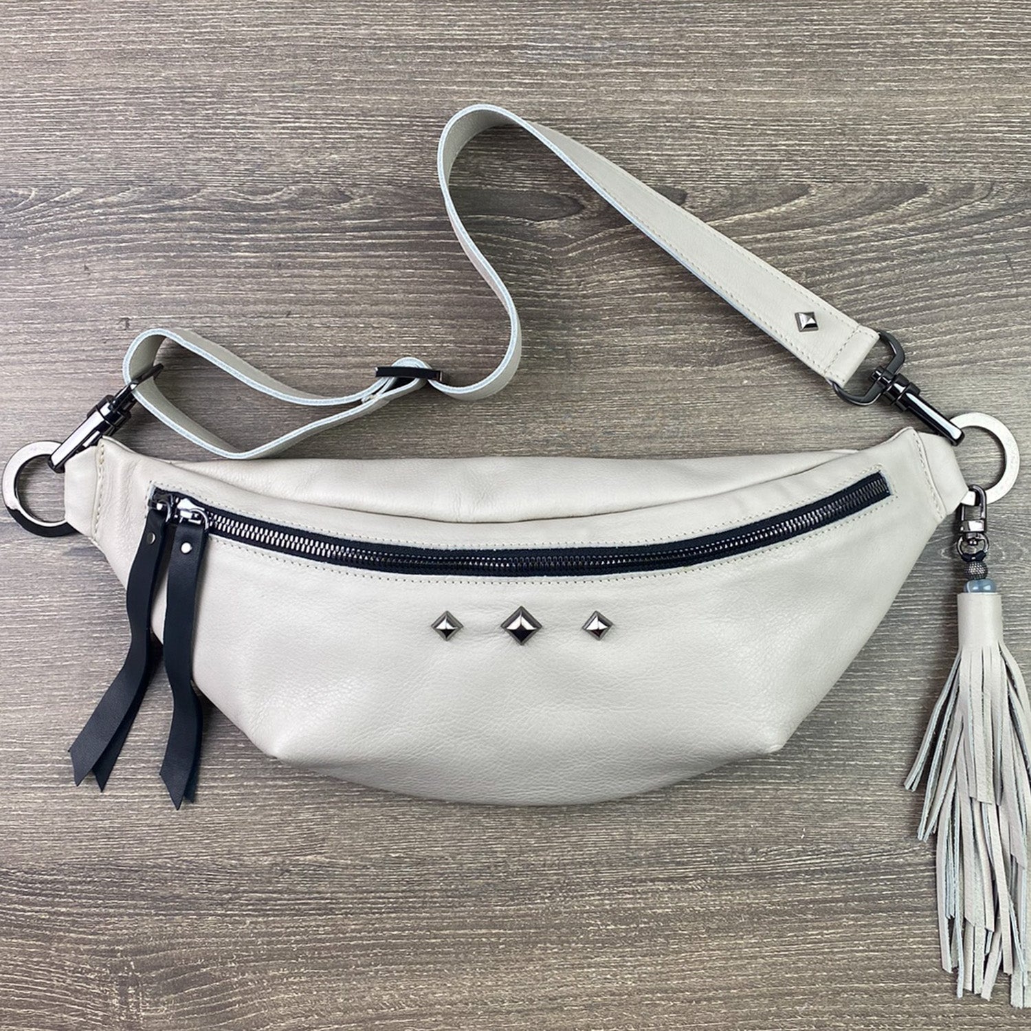 The STELLA - PUTTY cross body bag by evokewomen, featuring a beige leather design with black zippers, metal studs, and a decorative tassel, rests on a wooden surface.