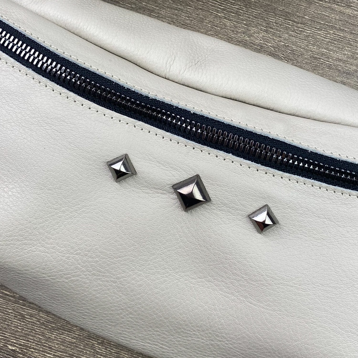 Close-up of the STELLA - PUTTY, a white leather cross body bag by evokewomen, featuring a zipper and three metallic pyramid studs.