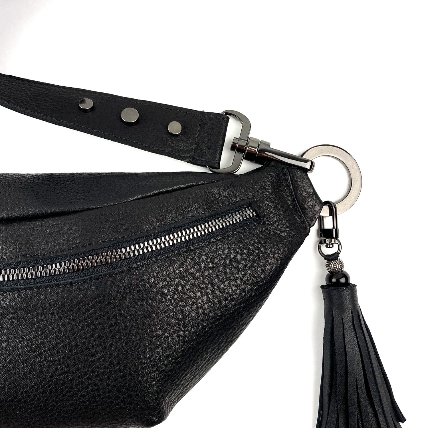 Close-up of the STELLA - BLACK cross body bag by evokewomen, showcasing its sleek black leather finish with a zippered pocket, metal hardware, and an elegantly attached tassel keychain on a ring.