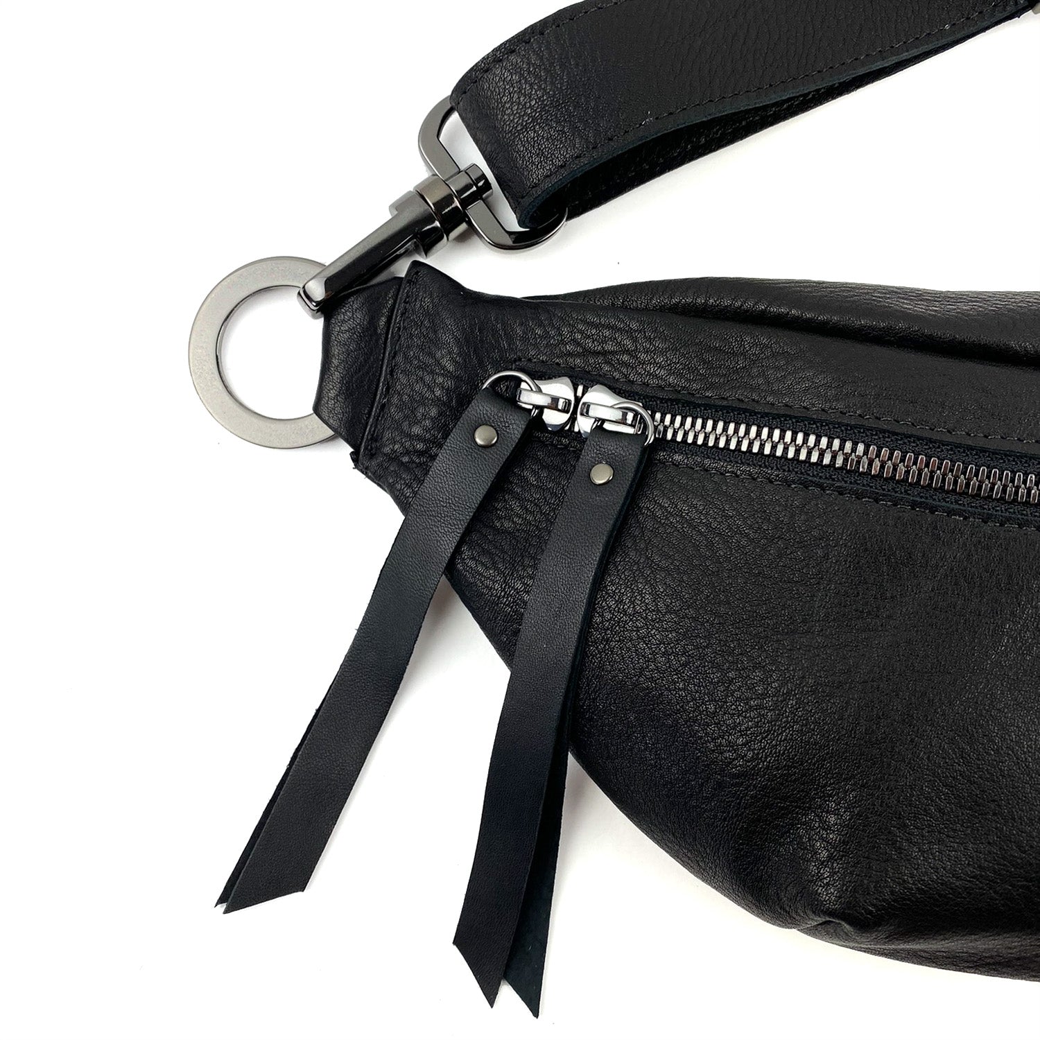 Close-up of the STELLA - BLACK by evokewomen, a sleek black leather sling bag featuring a metal zipper, two leather tassels, and a circular metal ring on the strap.