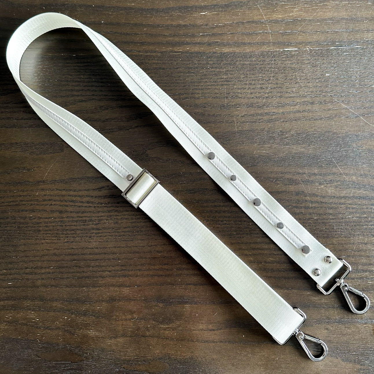 An off-white SHOULDER STRAP by evokewomen, featuring durable nylon webbing straps and metal clips, is laid flat on a wooden surface.