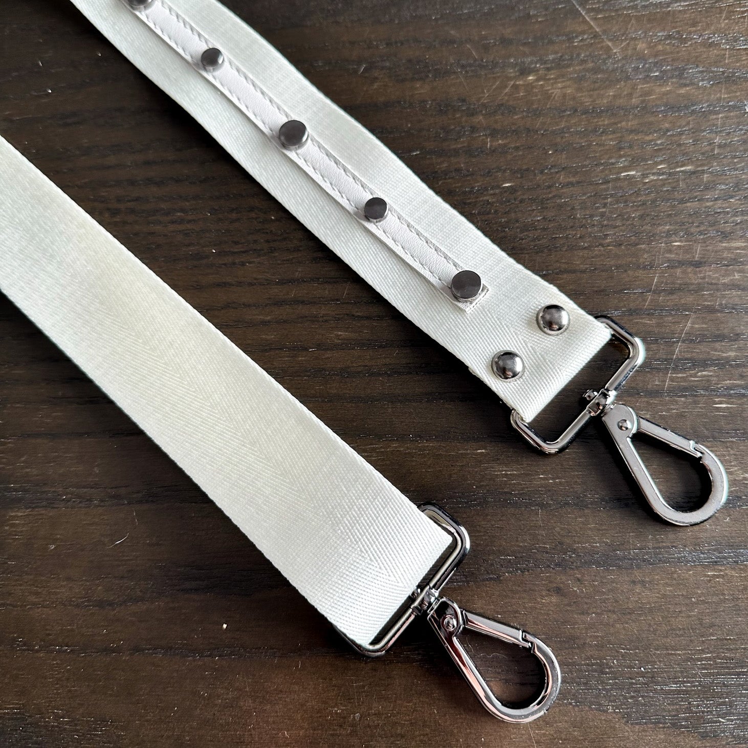 The SHOULDER STRAP - OFF WHITE by evokewomen is displayed on a wooden surface, featuring white nylon webbing with metal snap buttons and swivel hooks, highlighting its adjustable design for versatile use.