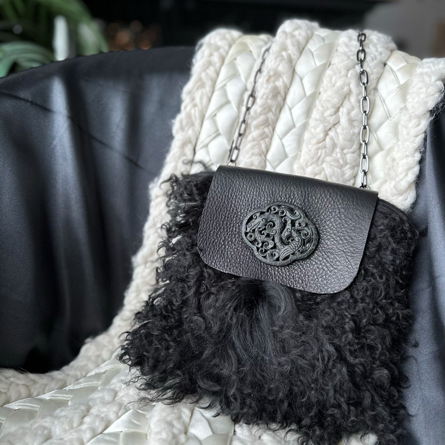 The MIA - BLACK by evokewomen, featuring a curly textured body and an ornate clasp, elegantly hangs like a chic shoulder bag on a cream knitted blanket draped over a black chair.
