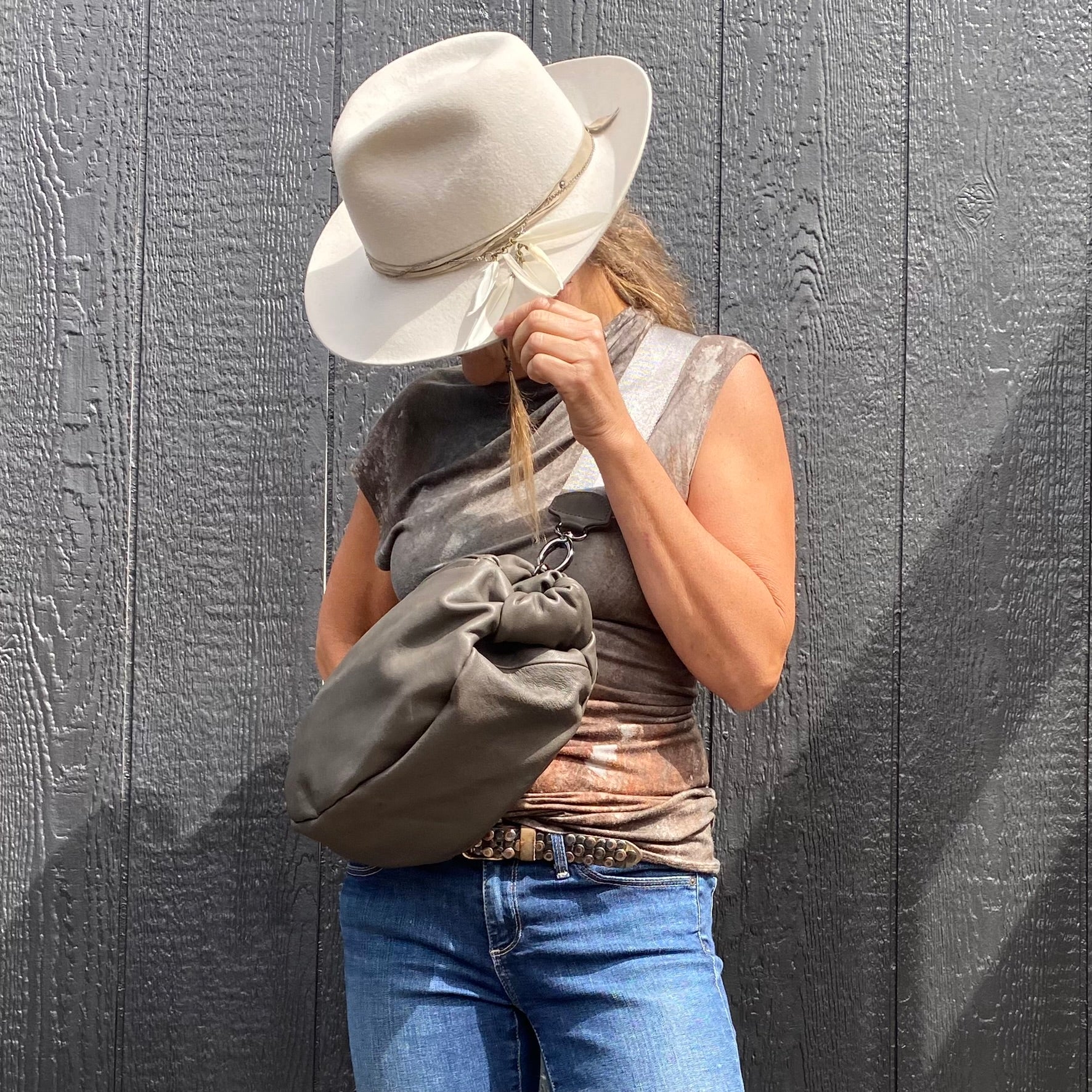 A person dressed in jeans and a sleeveless top holds the LULA - OLIVE-GREY leather crossbody bag by evokewomen, using a white cowboy hat to cover their face while standing in front of a textured gray wall.