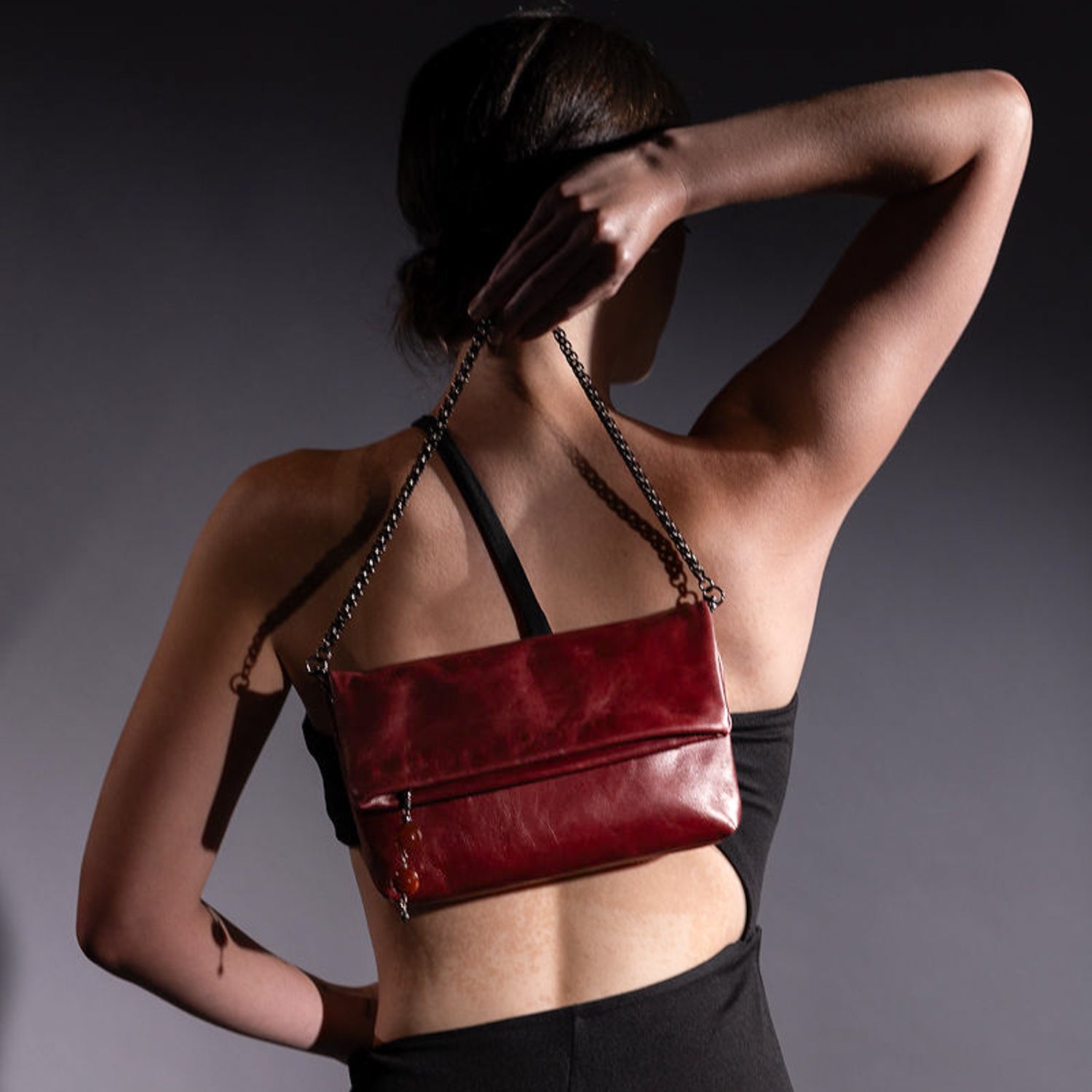 A person in a black top is elegantly posing against a dark background, holding the evokewomen Farah handbag in wine leather, featuring a distinctive snake spine chain strap, over their shoulder.