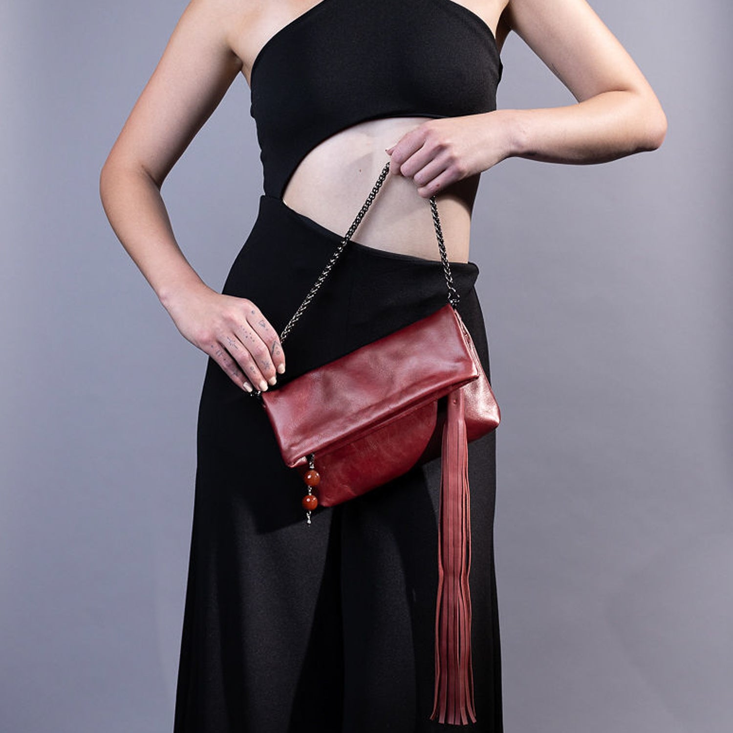 A person dressed in a black outfit holds the evokewomen TASSEL XXL in wine, showcasing its standout design feature: an oversized tassel and chain, creating a unique look as it rests in front of their waist.