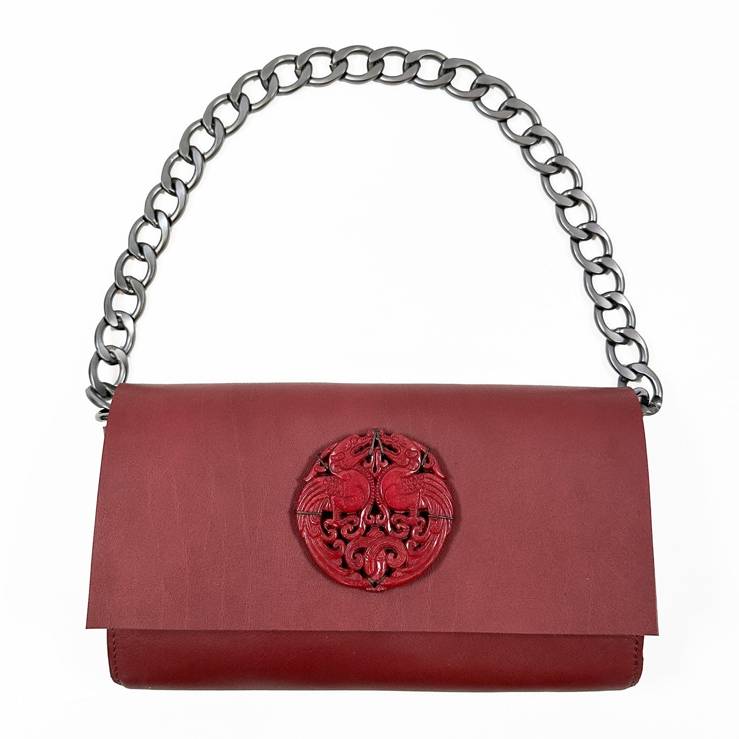 Introducing the BAGUETTE - WINE by evokewomen, an opulent red handbag made from Italian leather. It features a chic chain strap and a distinctive decorative emblem on the flap, evoking the elegance of a vintage jade amulet.