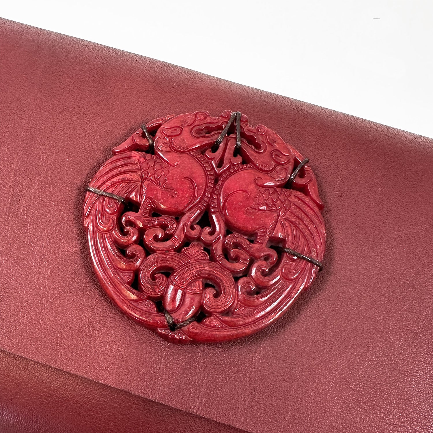 Introducing the BAGUETTE - WINE by evokewomen, a stunning piece crafted from exquisite Italian leather. This round red design features an intricate carving of two mirrored phoenixes, echoing the elegance of a vintage jade amulet with its detailed swirling patterns.