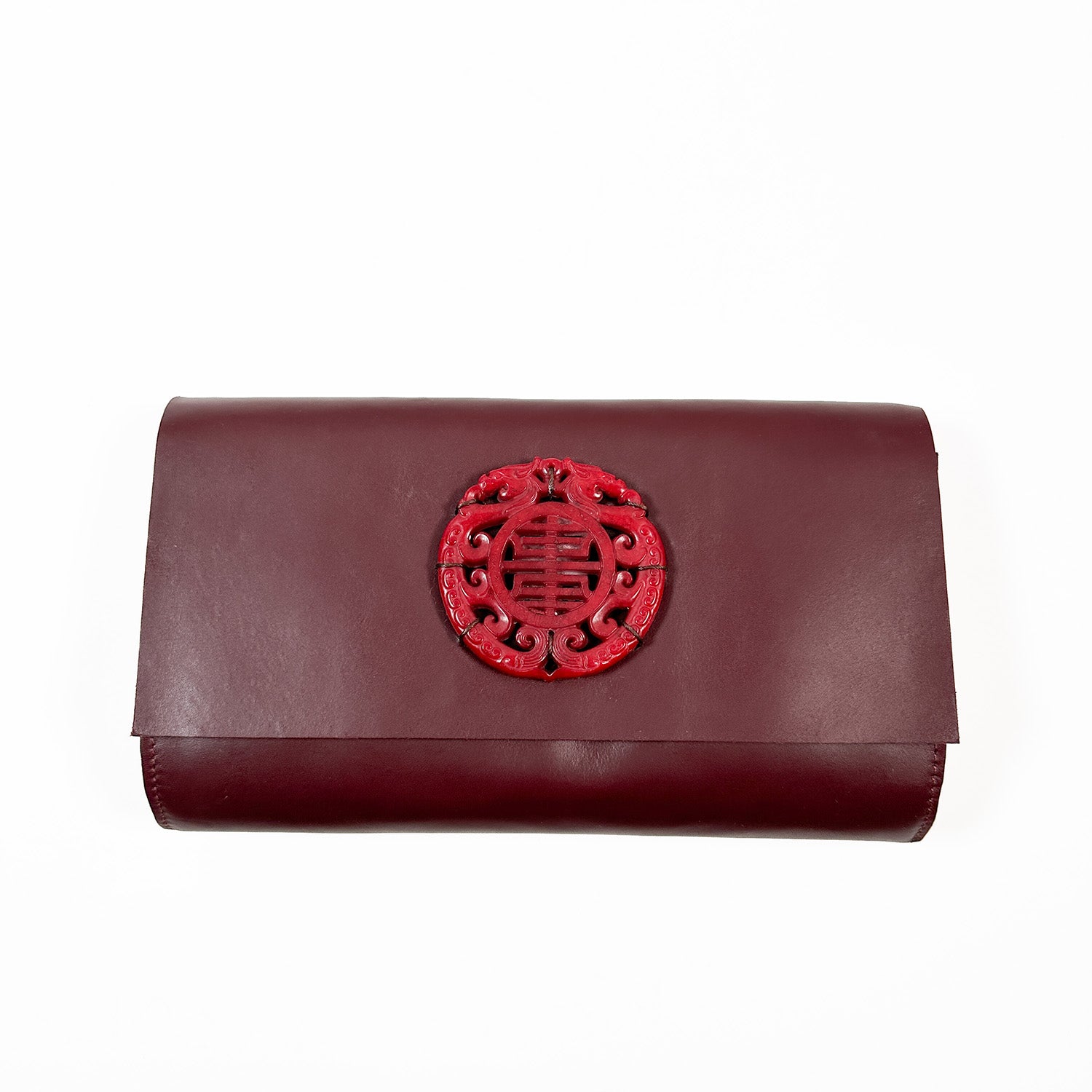 The BAGUETTE - BURGUNDY by evokewomen is fashioned from luxurious Italian leather in a rich maroon hue, adorned with a bold red circular emblem showcasing intricate cut-out designs on the front flap.