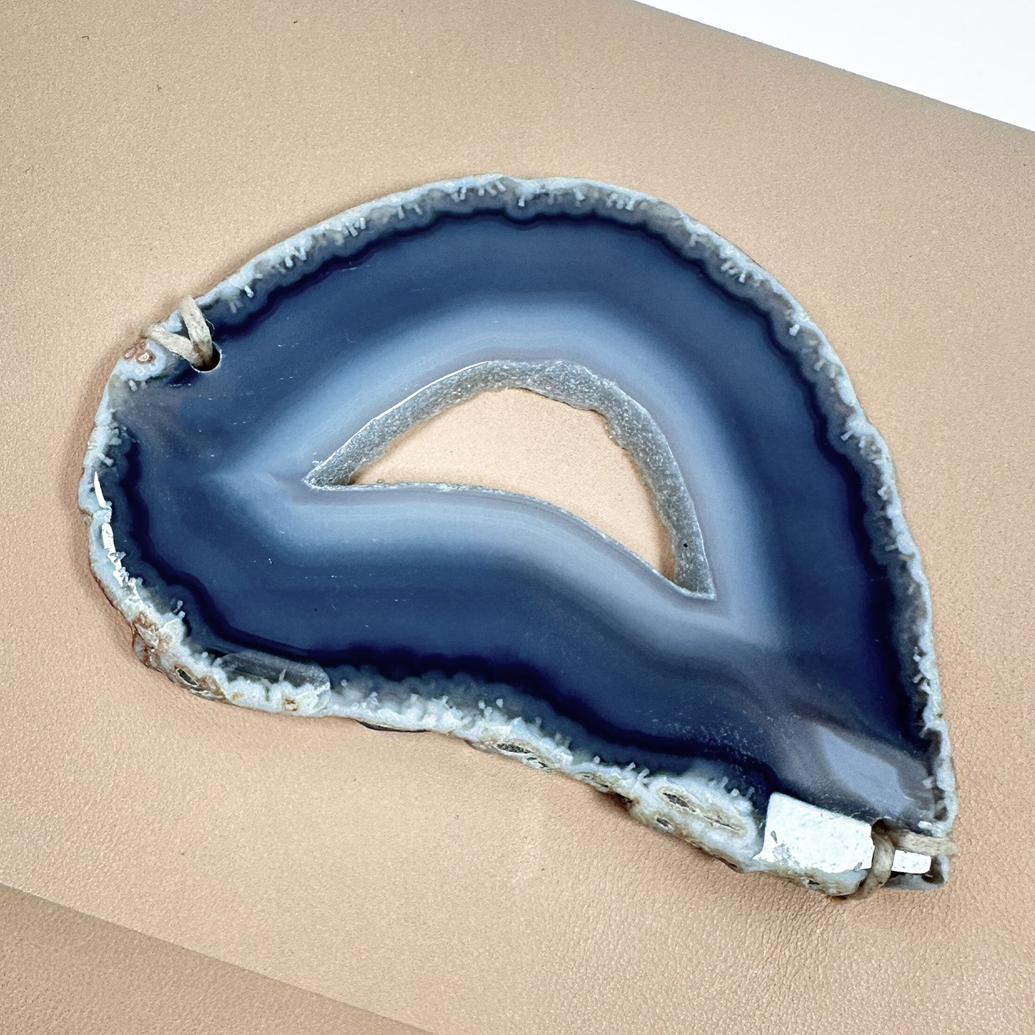 Close-up of a teardrop-shaped blue/grey agate slice with natural banding and an open center, displayed on a smooth beige leather surface.