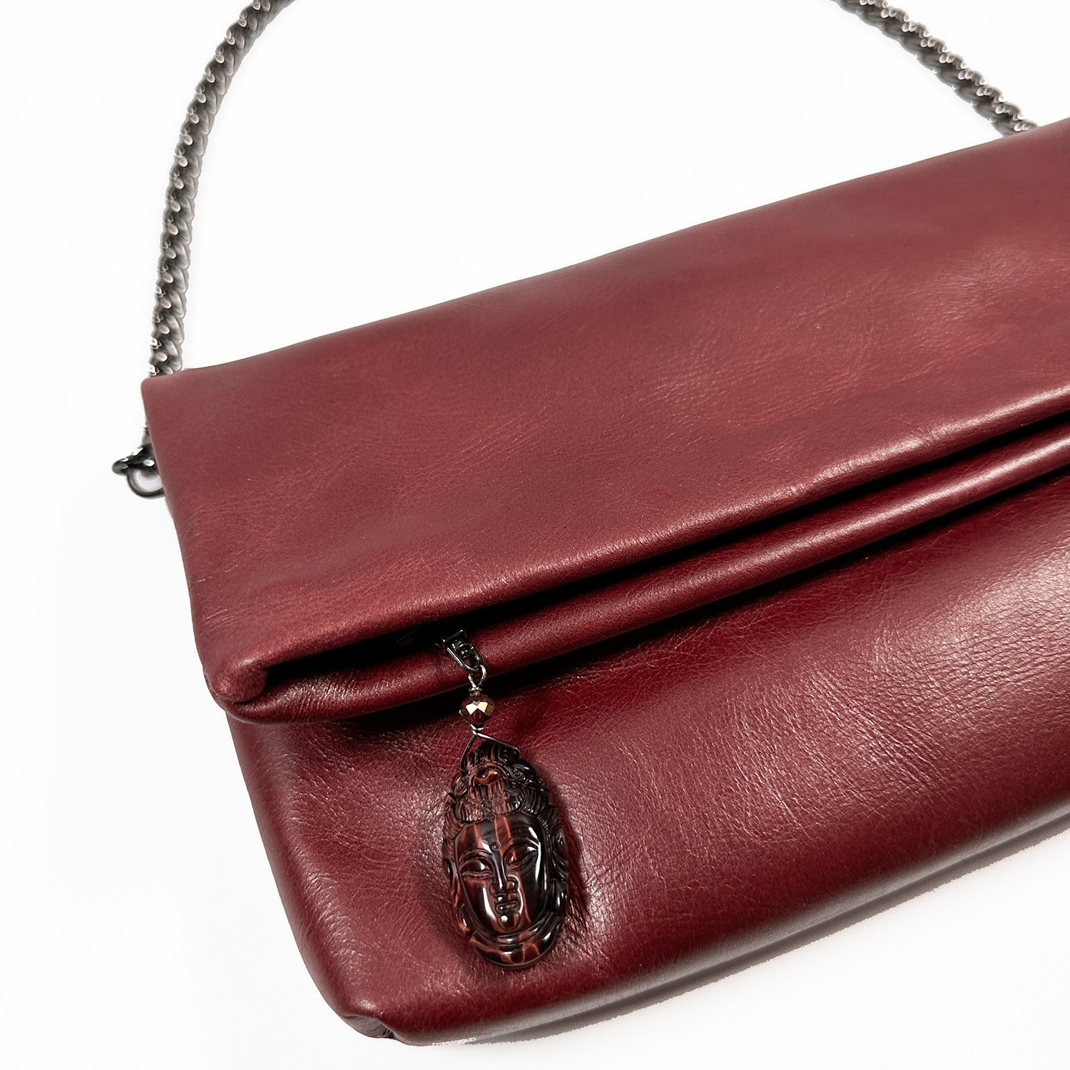 A wine-colored Farah clutch by evokewomen, crafted from leather and featuring a Snake Spine Chain strap with a decorative face pendant on the flap.