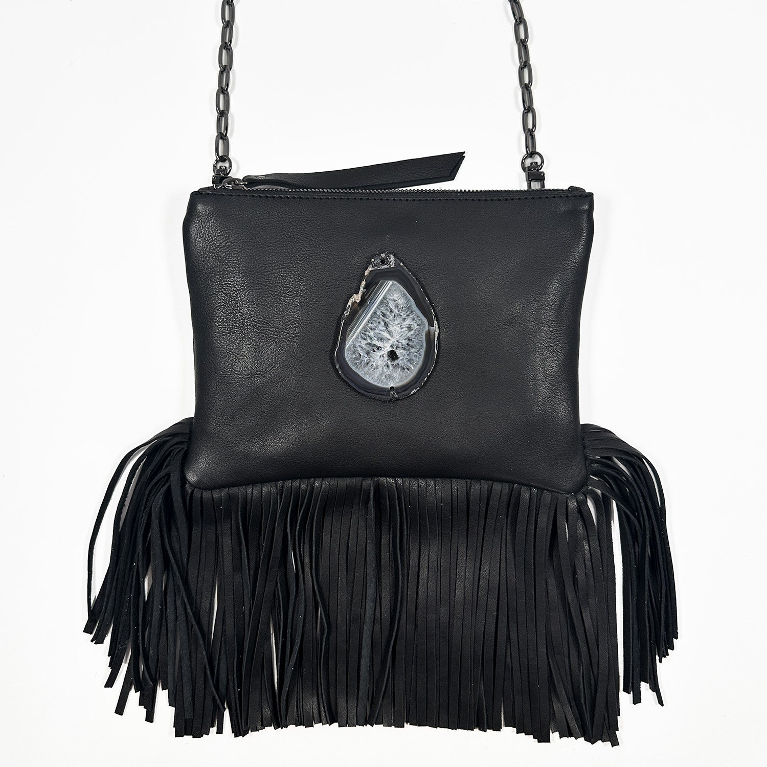 Black cross-body bag with zipper closure, fringes at bottom and teardrop shaped black edged agate stone with natural crystal formation on white background. Gunmetal cross-body chain. 