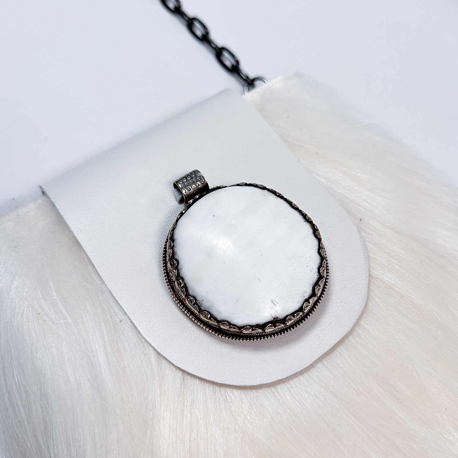 The MIA - WHITE pendant by evokewomen is showcased on a white furry surface, featuring a captivating round silver design with a white stone, reminiscent of an exquisite Tibetan bone amulet, and is elegantly attached to a black chain.