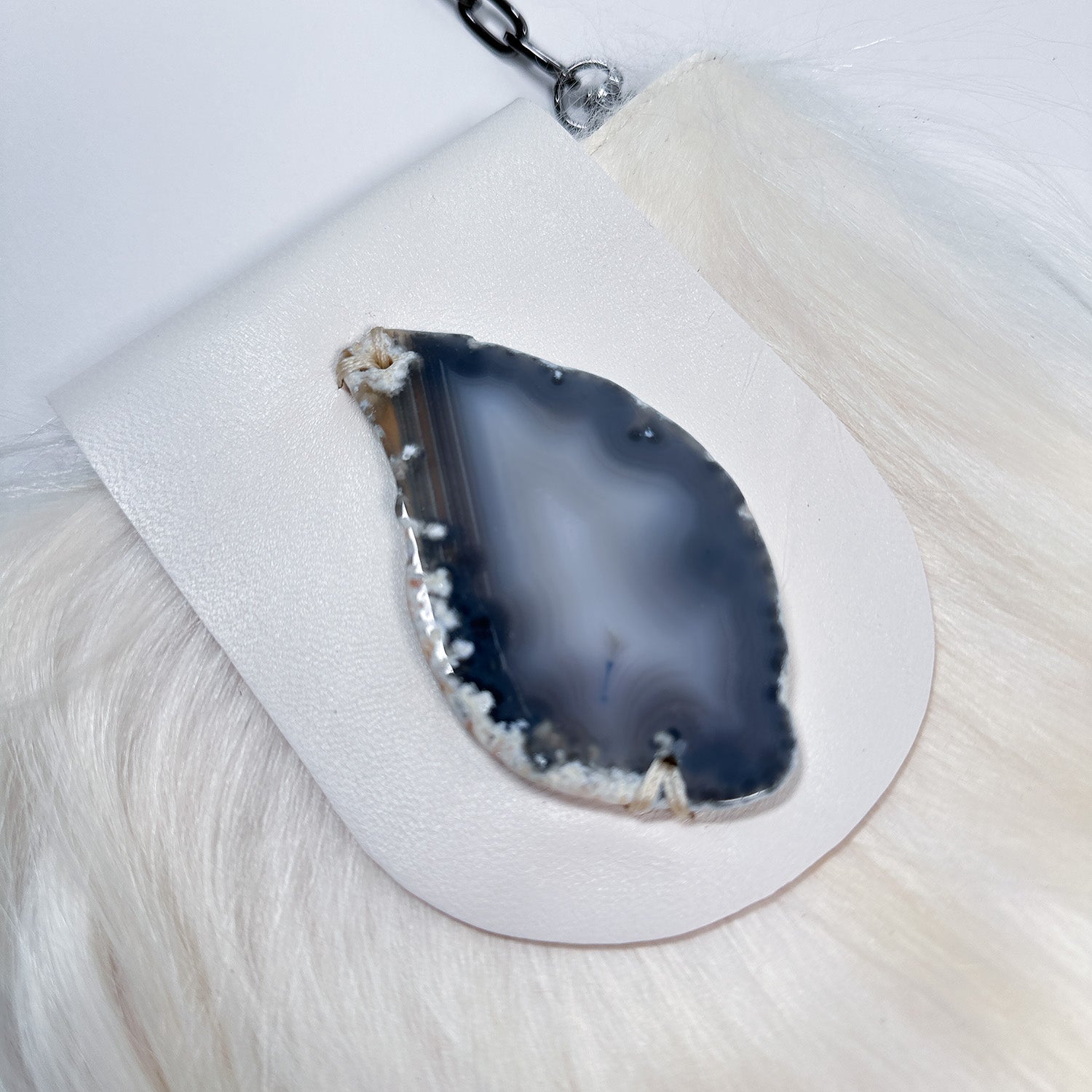 A close-up of the MIA - WHITE crossbody bag by evokewomen, featuring a smooth, teardrop shape with a translucent blue and gray pattern, set against a white and furry background.