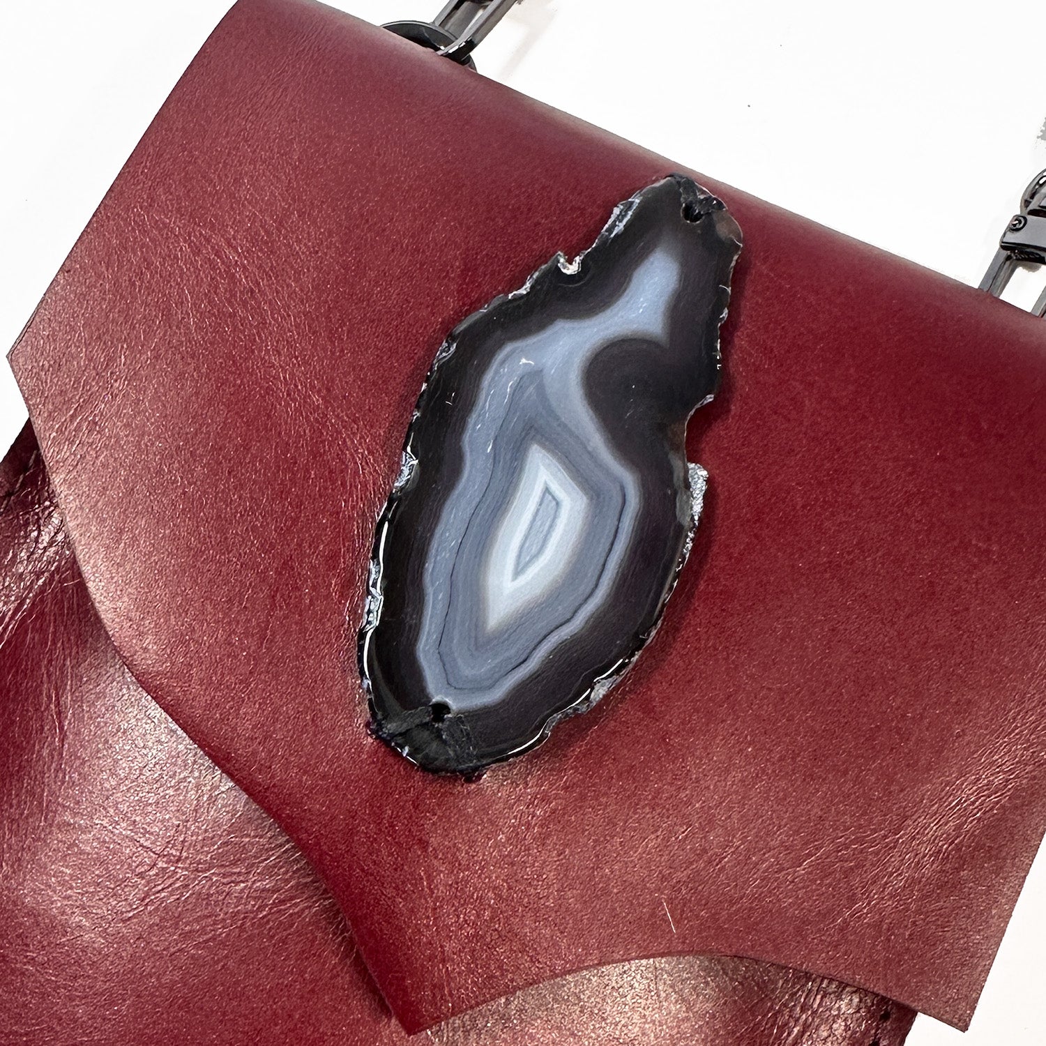 An essential for festivals, the PAULINA - wine cross body bag by evokewomen is crafted from maroon Italian leather and showcases a striking black and white geode slice embellishment on the flap.