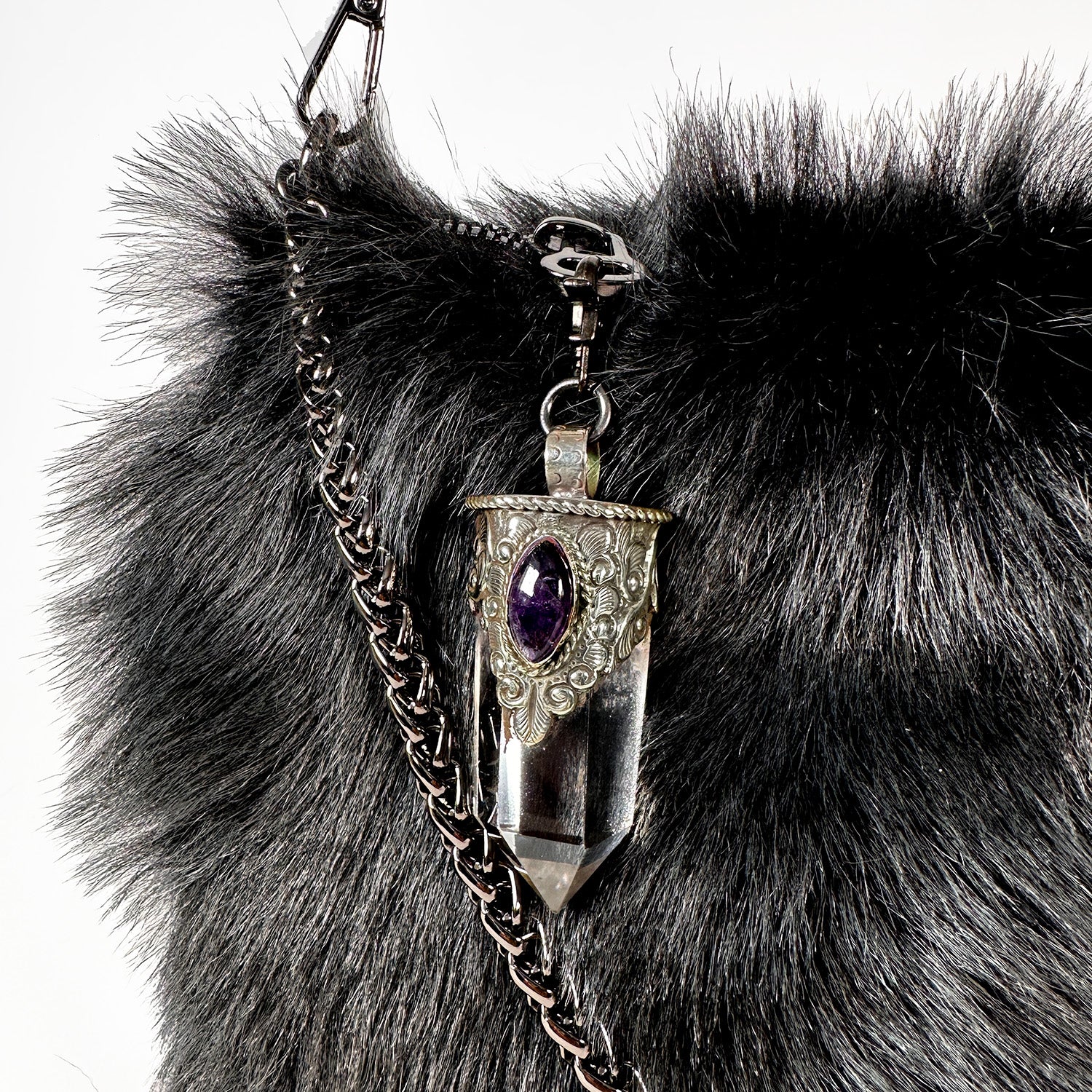 A close-up of the POPPY - BLACK LIGHT TIPPED bag by evokewomen reveals a silver decorative pendant adorned with an amethyst stone and intricate metalwork. This chic Toscana Lamb Fur crossbody bag enhances your style with its unique Tibetan crystal rock pendant charm.